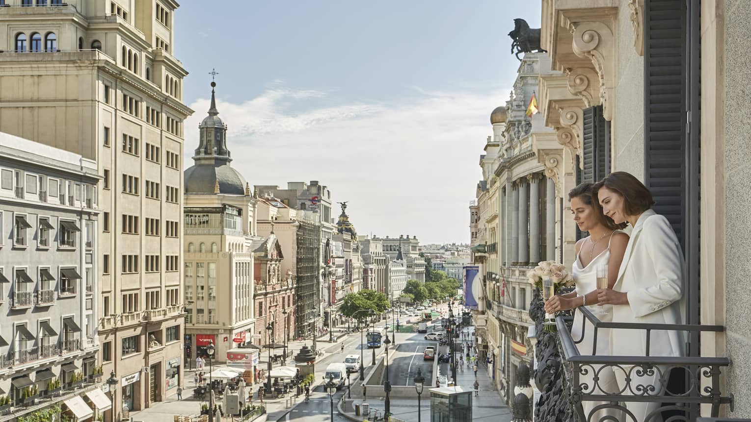 Wedding couple in elegant attire stand on a balcony overlooking a bustling city street lined with historic buildings, enjoying a quiet moment with champagne.