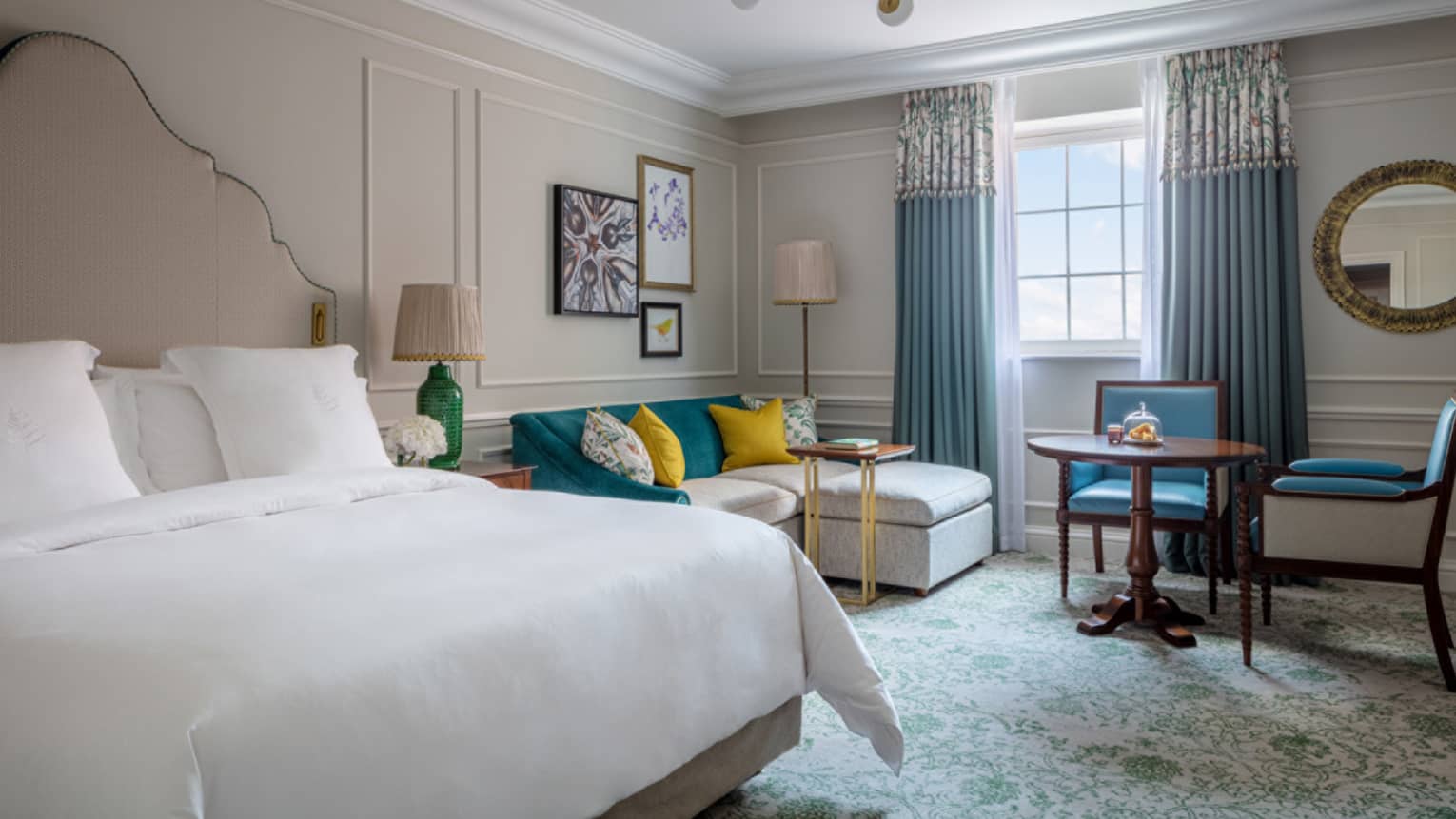 Guest room with king bed and white linens, cream sectional sofa with colourful pillows, small round table and two chairs, window with blue drapes