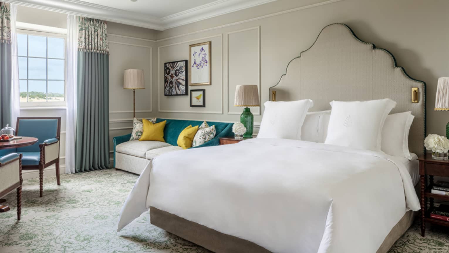 Guest room with king bed and white linens, cream couch with colourful pillows, small round table and two chairs, window with blue drapes