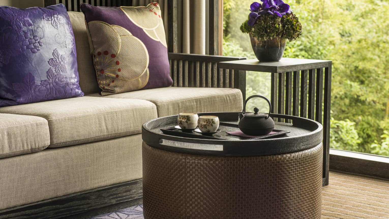 Close-up of beige sofa with purple pillows, round coffee table, garden view out window