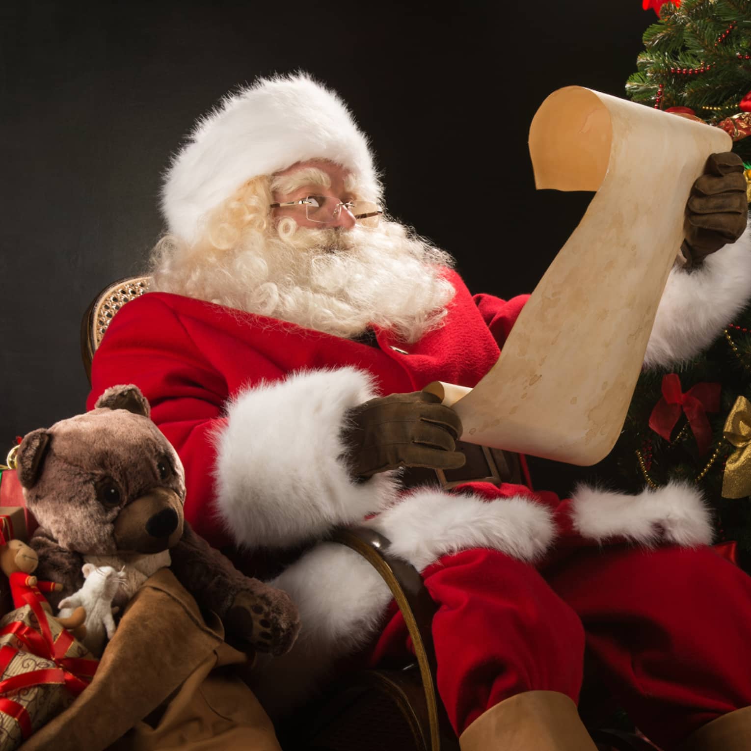 Santa Claus seated in large rocker holding a scroll, flanked by decorated Christmas tree with gifts and overflowing gift bag