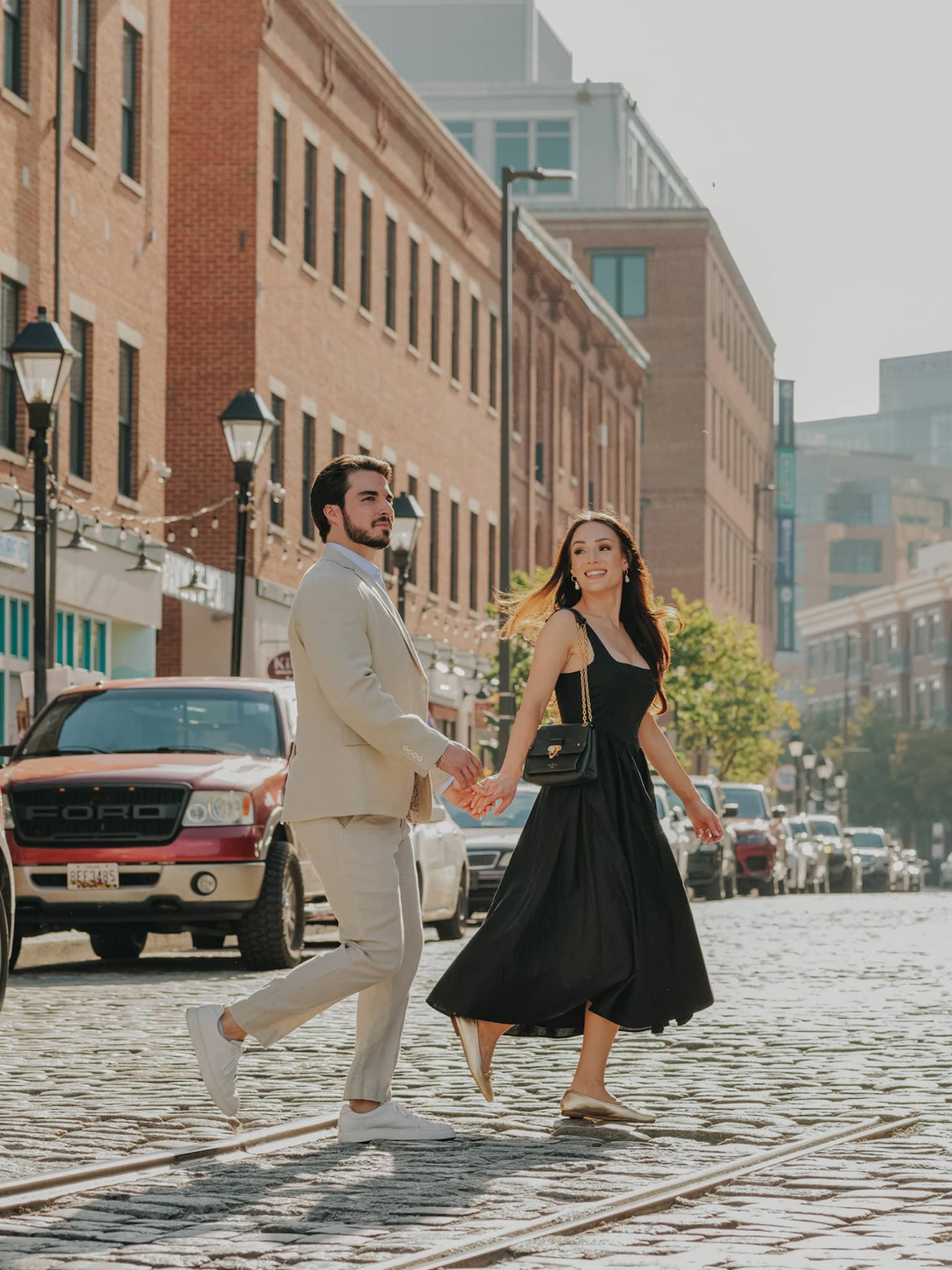 A smiling, well-dressed couple cross a cobbled street lined with stores in red-brick buildings, streetlights and parked cars.