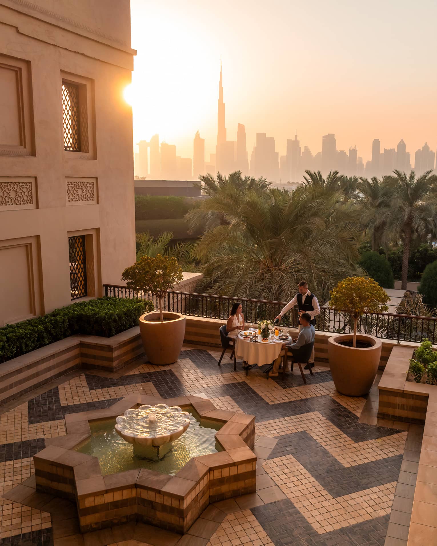 Table set for two on an open-air rooftop terrace with a chevron tiled floor and the sun rising over a city skyline in the near distance