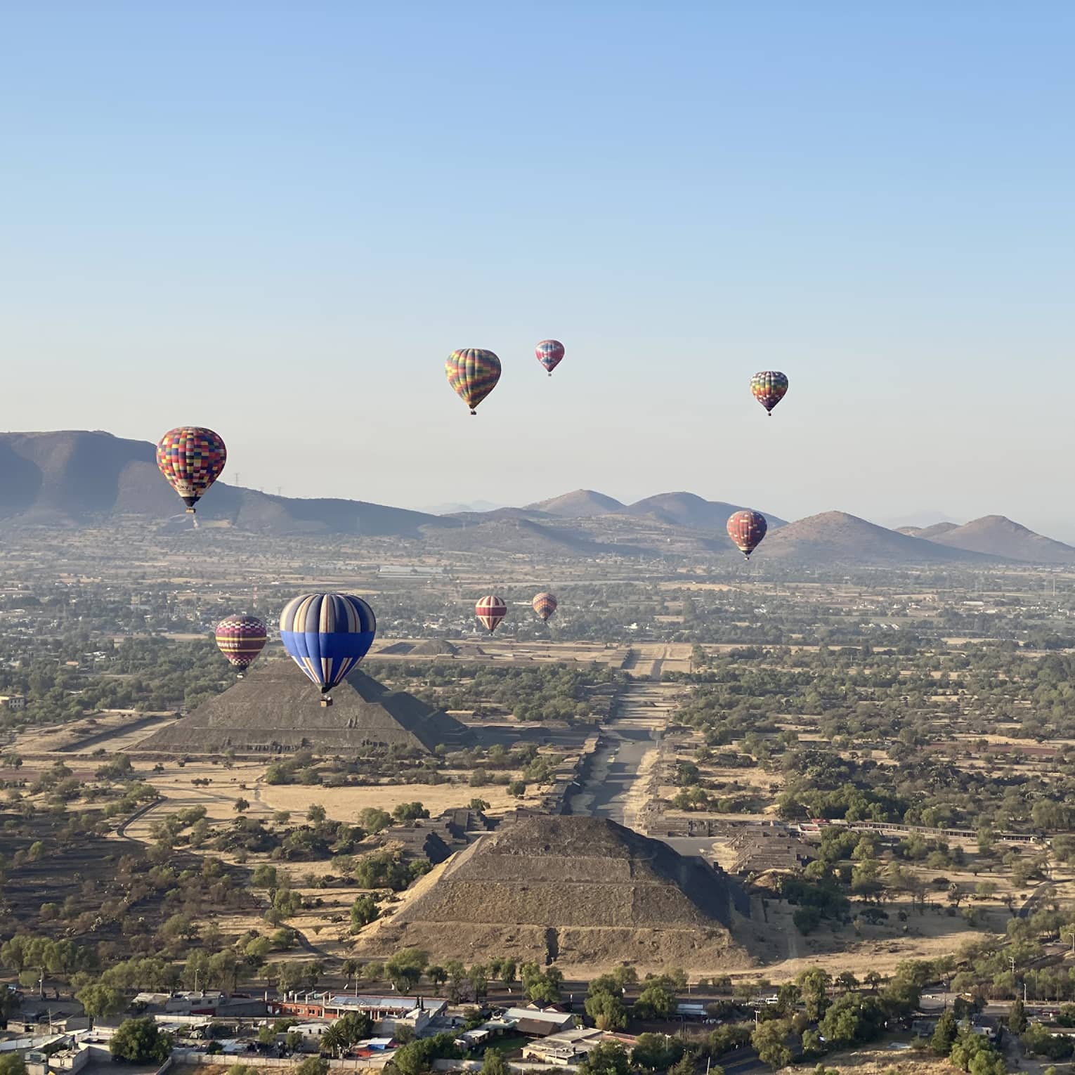 Hot-air balloons floating over old temples in a dessert covered in shrubs.