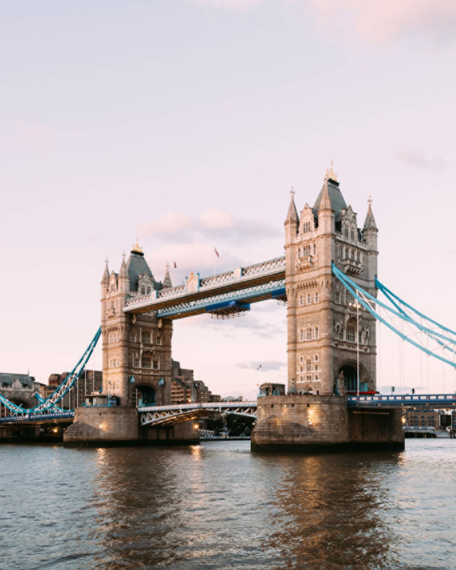 The iconic Tower Bridge of London spanning the dark Thames River in the foreground against a pale, dusky sky.