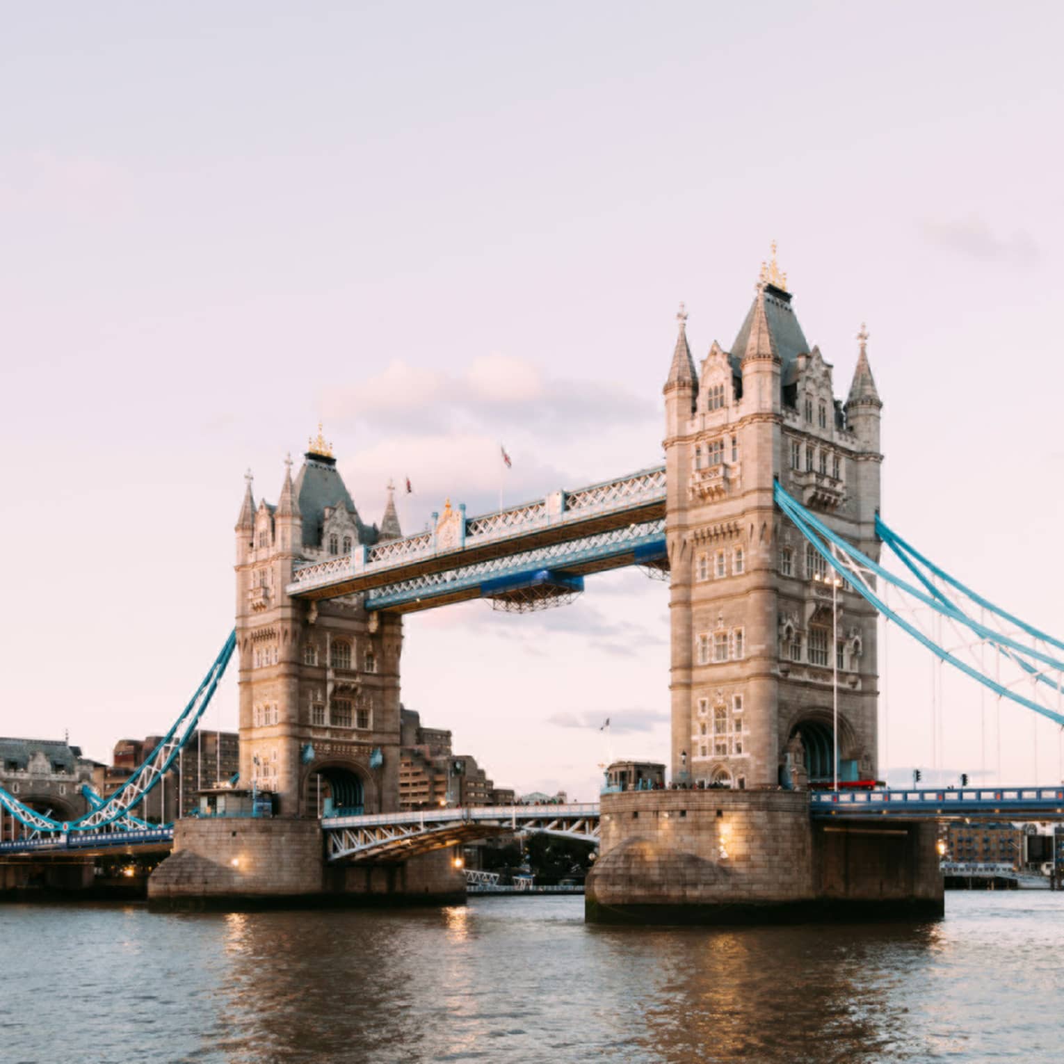 The iconic Tower Bridge of London spanning the dark Thames River in the foreground against a pale, dusky sky.