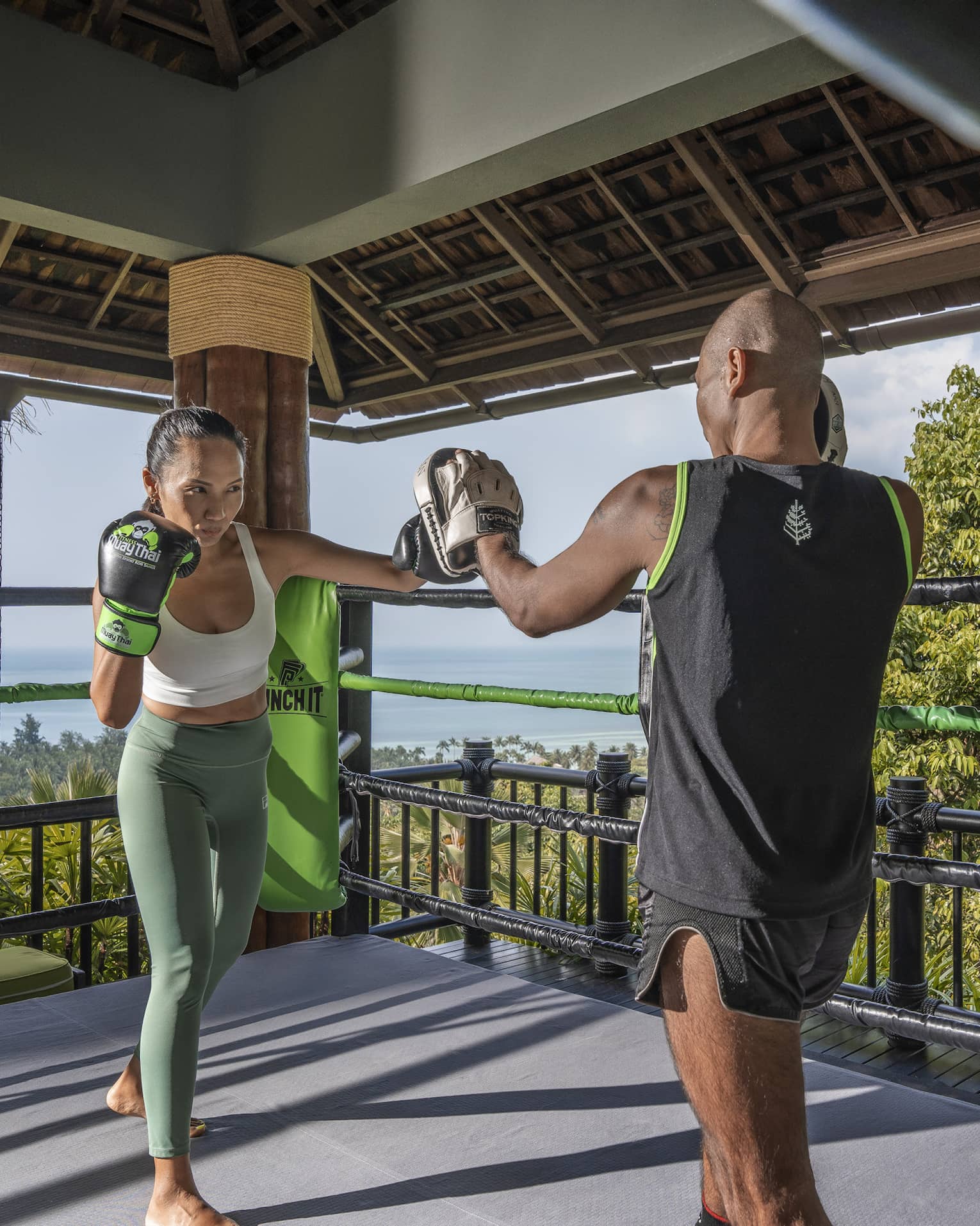 Two people practise Muay Thai in an outdoor boxing ring nestled in the treetops and overlooking the forest and the ocean.