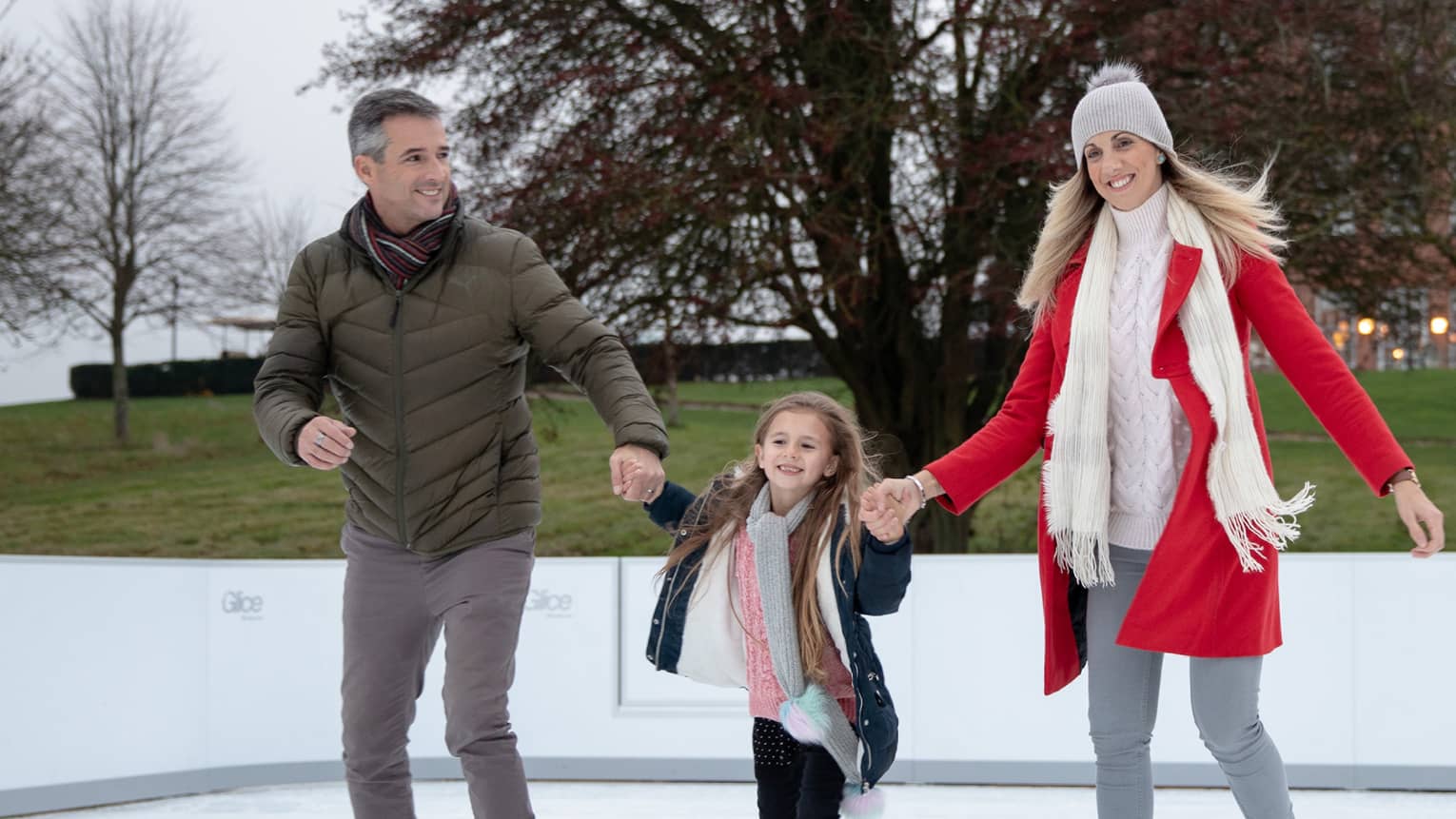 Man and woman holding hands of young girl while ice skating on outdoor rink