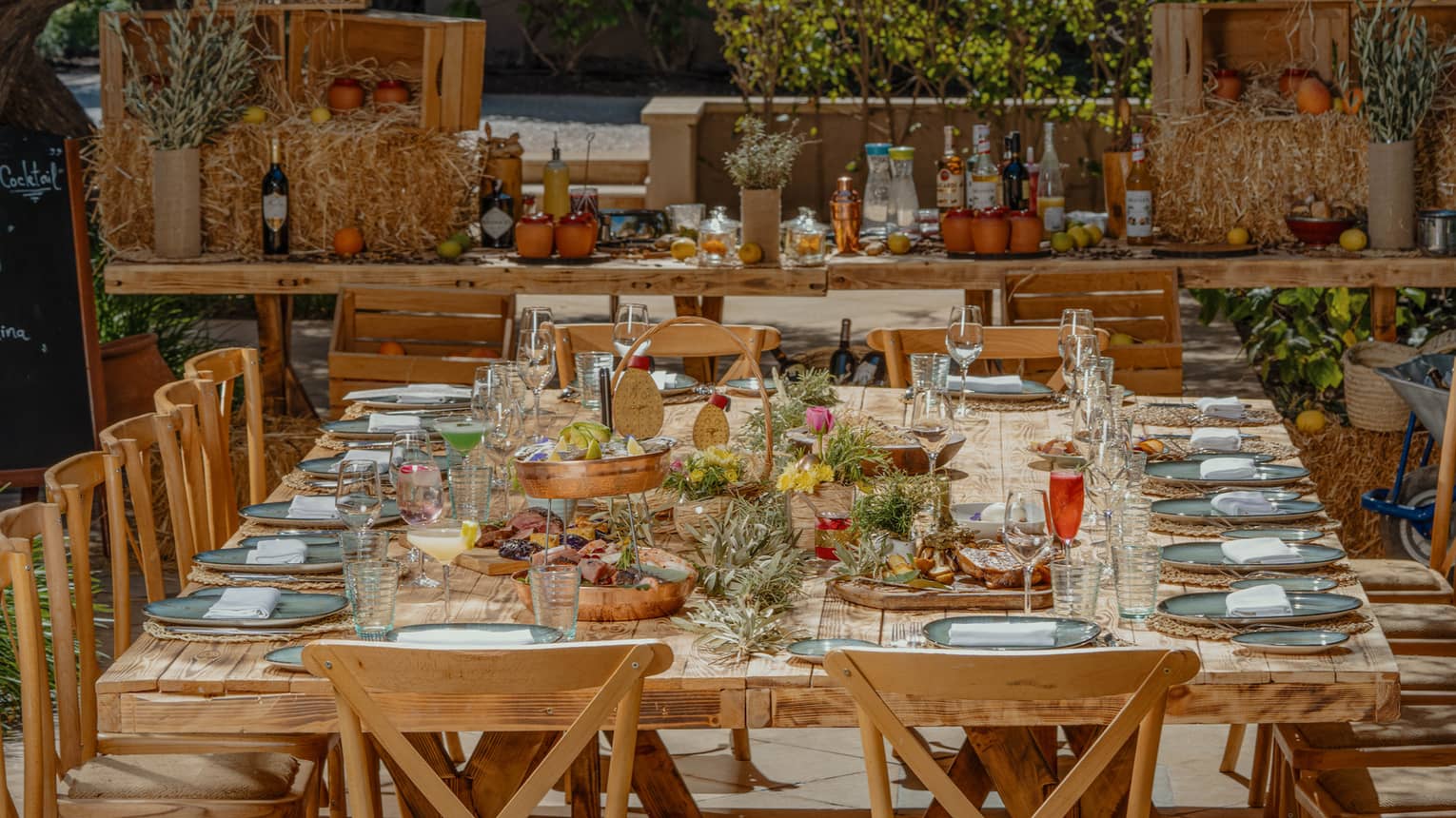 A large wooden dining table surrounded by cross back chairs and topped with plates, glasses and colorful floral arrangements sits on a patio covered with lush greenery