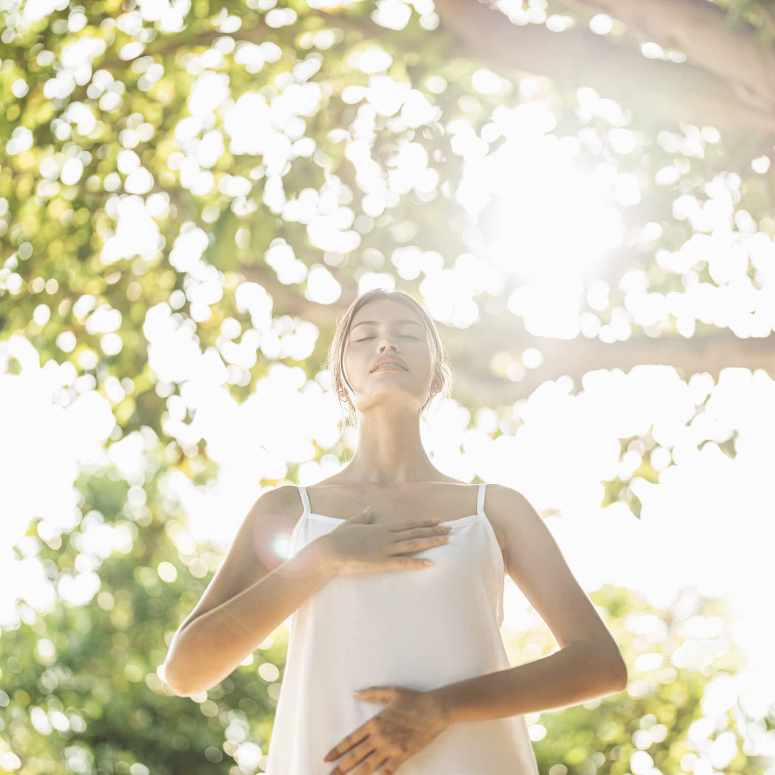 Person practicing mindful breathing in a wellness haven, standing under sunlit trees with hands on chest and abdomen