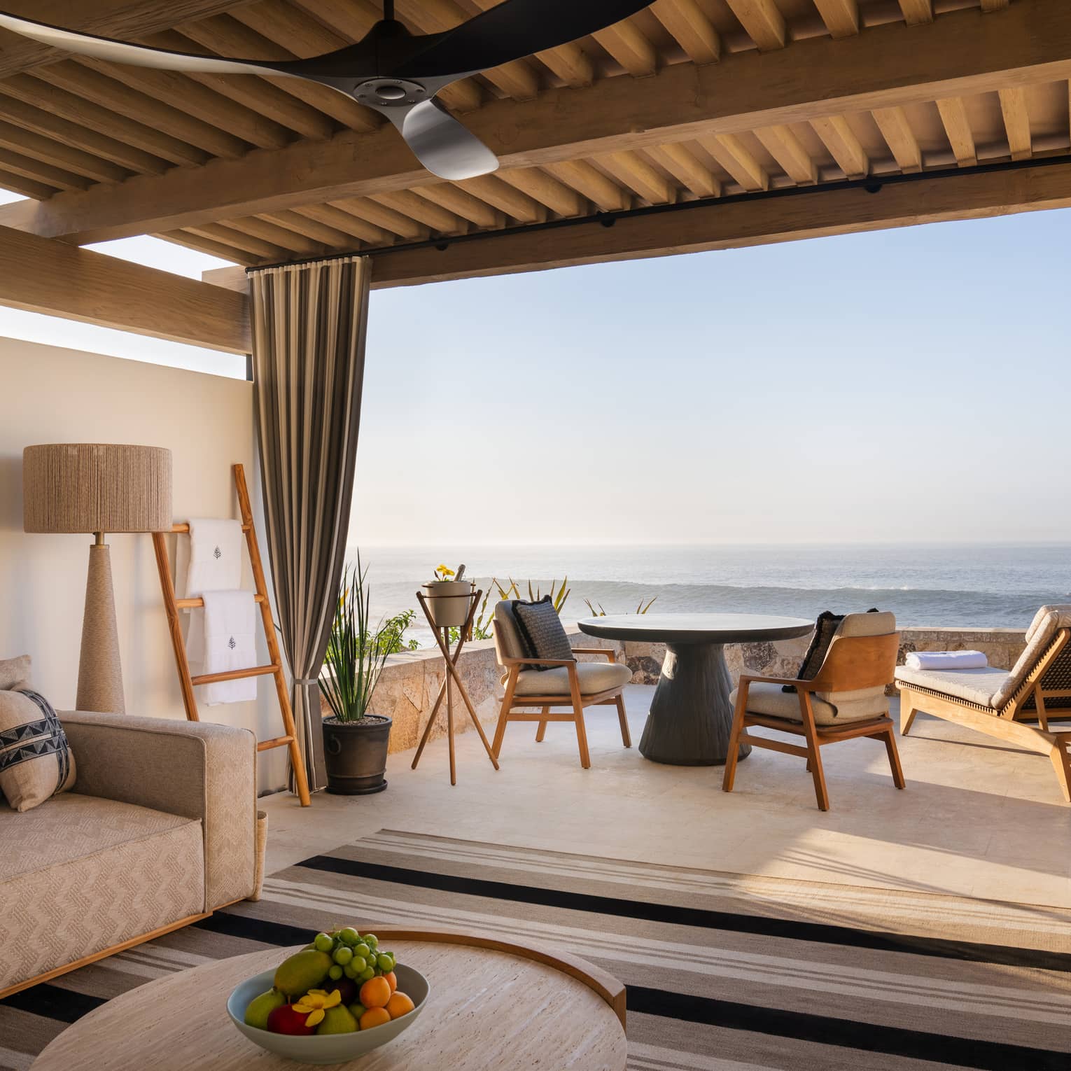 Oceanfront cabana with beige sofa, lamp and round wood coffee table under a roof with a round table and chairs and two lounge chairs out on the open-air patio overlooking the ocean
