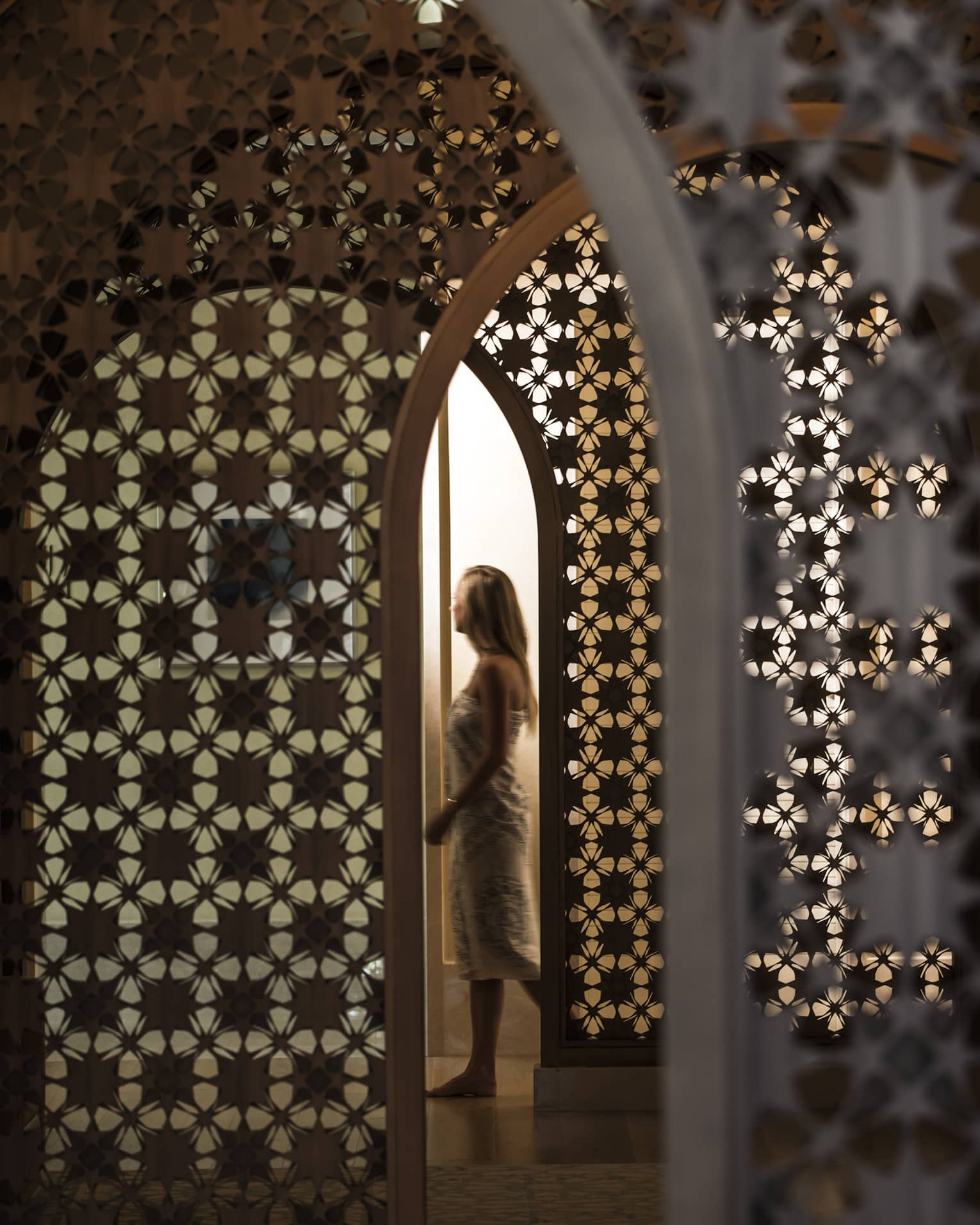 Woman in white wrap walks through dimly-lit decorative entranceway in hall