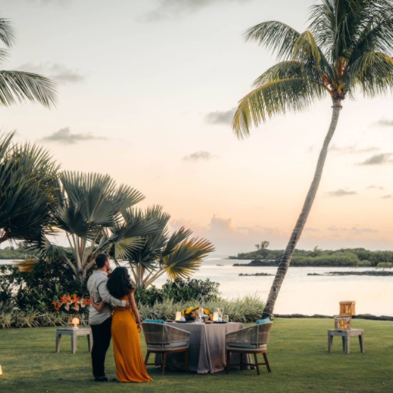 A couple stand on the shore, arms around each other, gazing toward the ocean; set table with two chairs beside swaying palms.