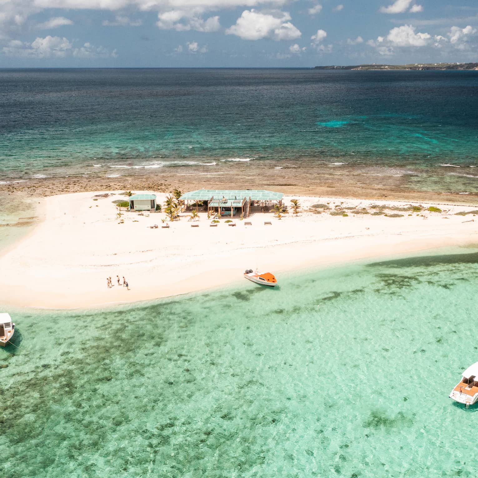 A small sandy island surrounded by crystal clear water and a few boats