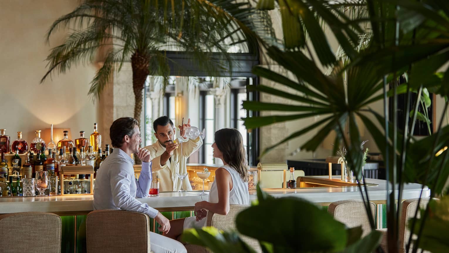 Mixologist pours cocktail for couple at small bar by potted palm trees