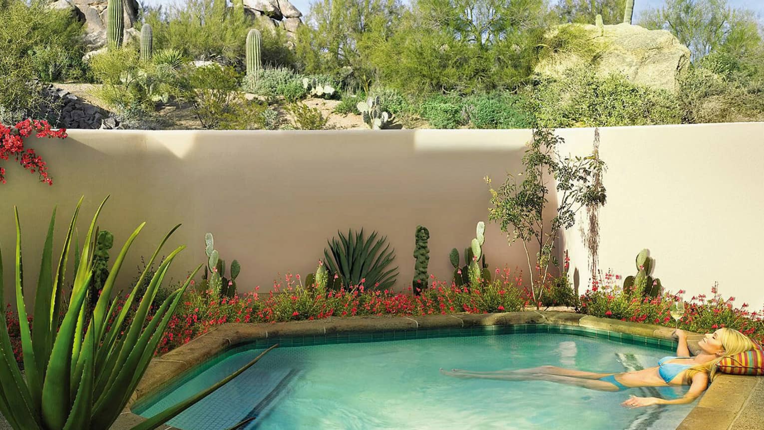 Woman wearing blue bikini wades in plunge pool with glass of white wine, cactus plants under white wall