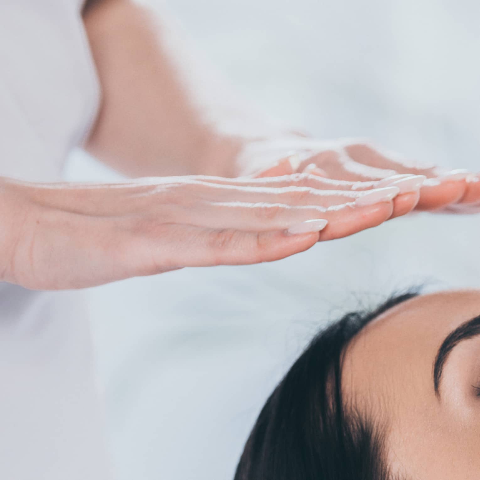 Woman with eyes closed lays on rolled up towel on spa table under therapists' hovering hands