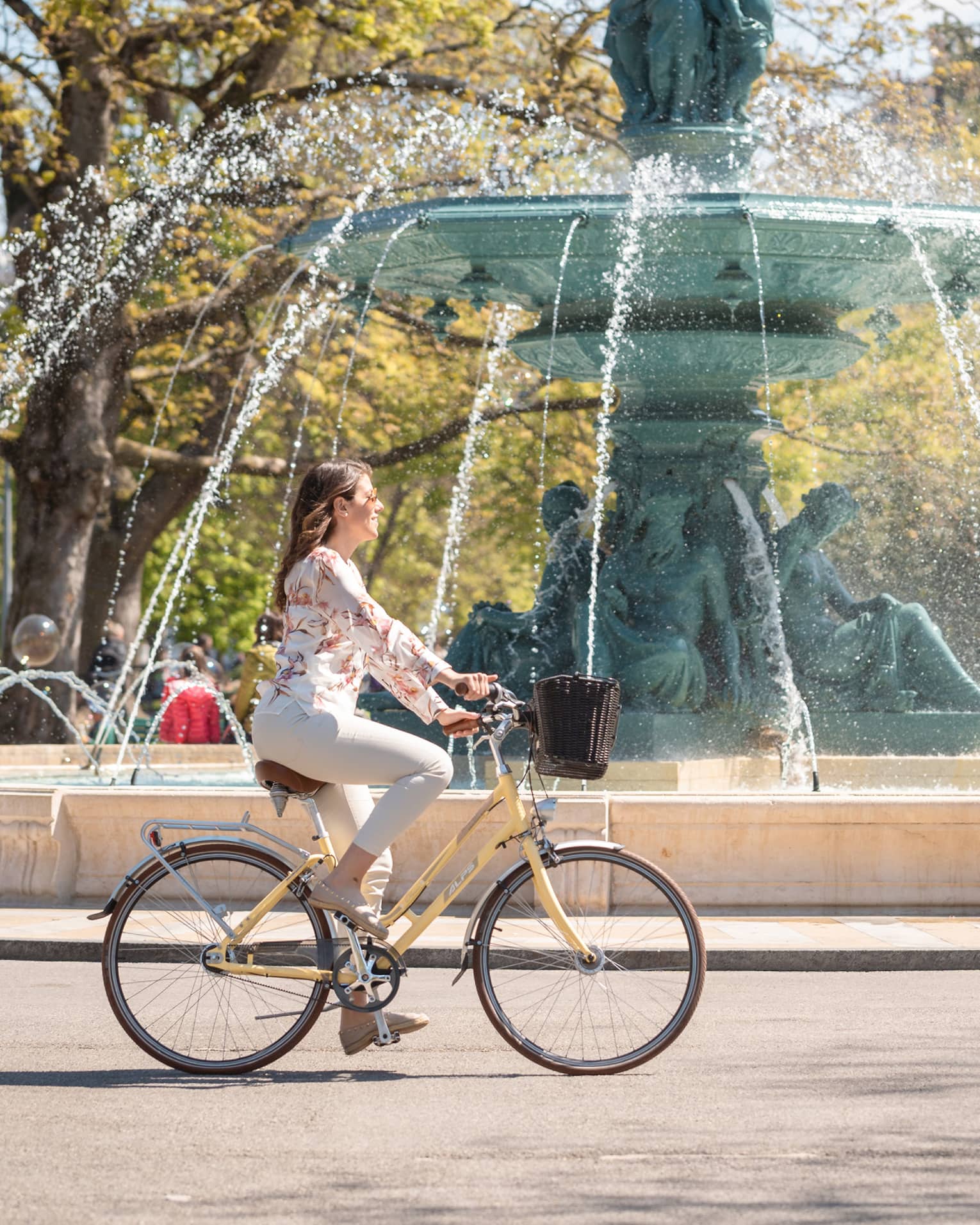 Side view of a guest on an old-style bike with a basket whizzing by a majestic fountain with a spout made from green statues.