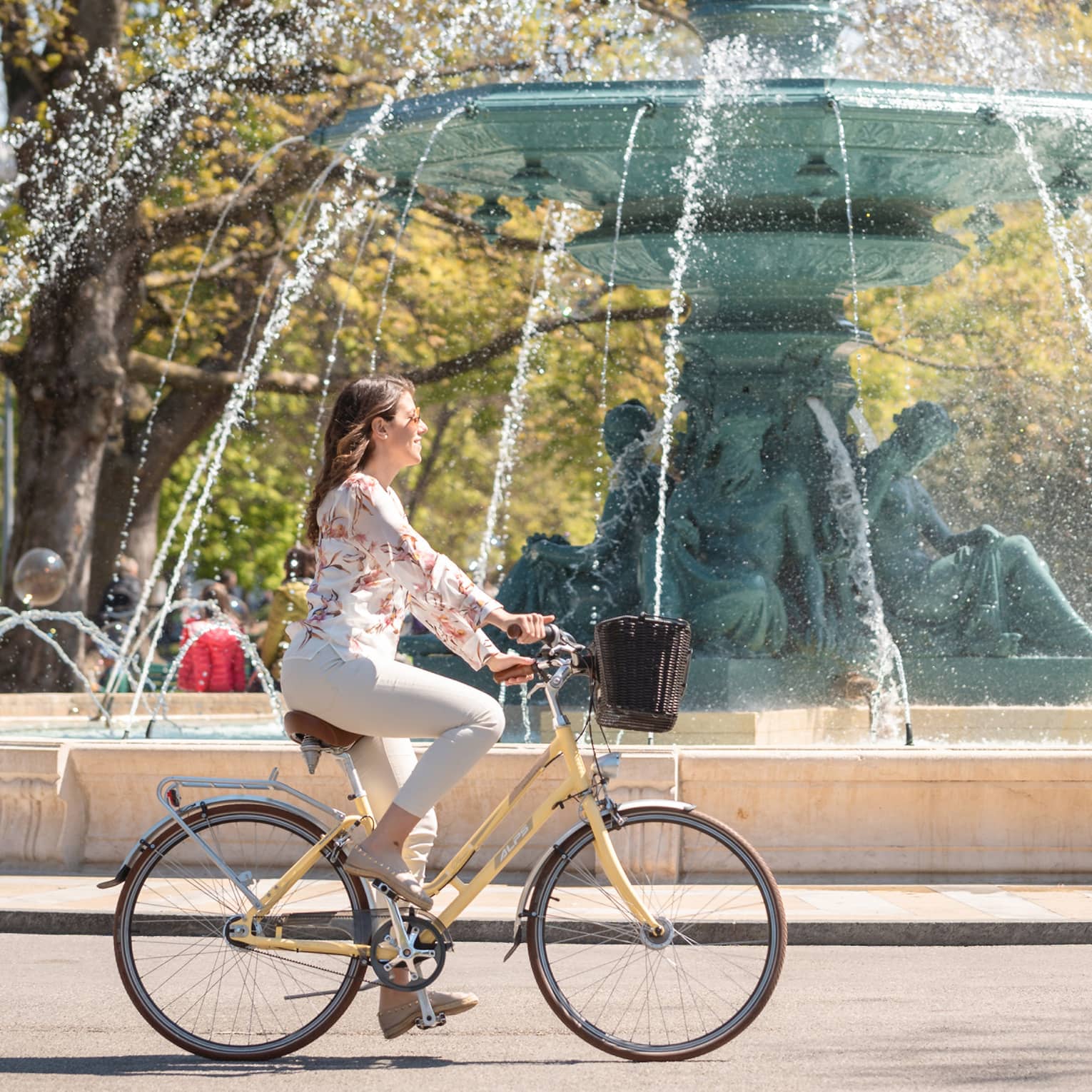 Side view of a guest on an old-style bike with a basket whizzing by a majestic fountain with a spout made from green statues.