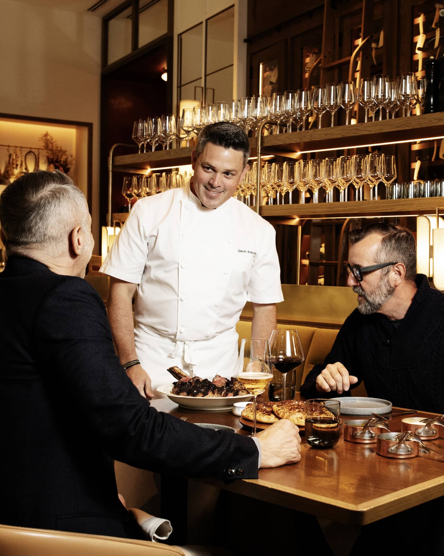 Two guests sitting at a dining table in a restaurant while the chef stands near the table talking to them.