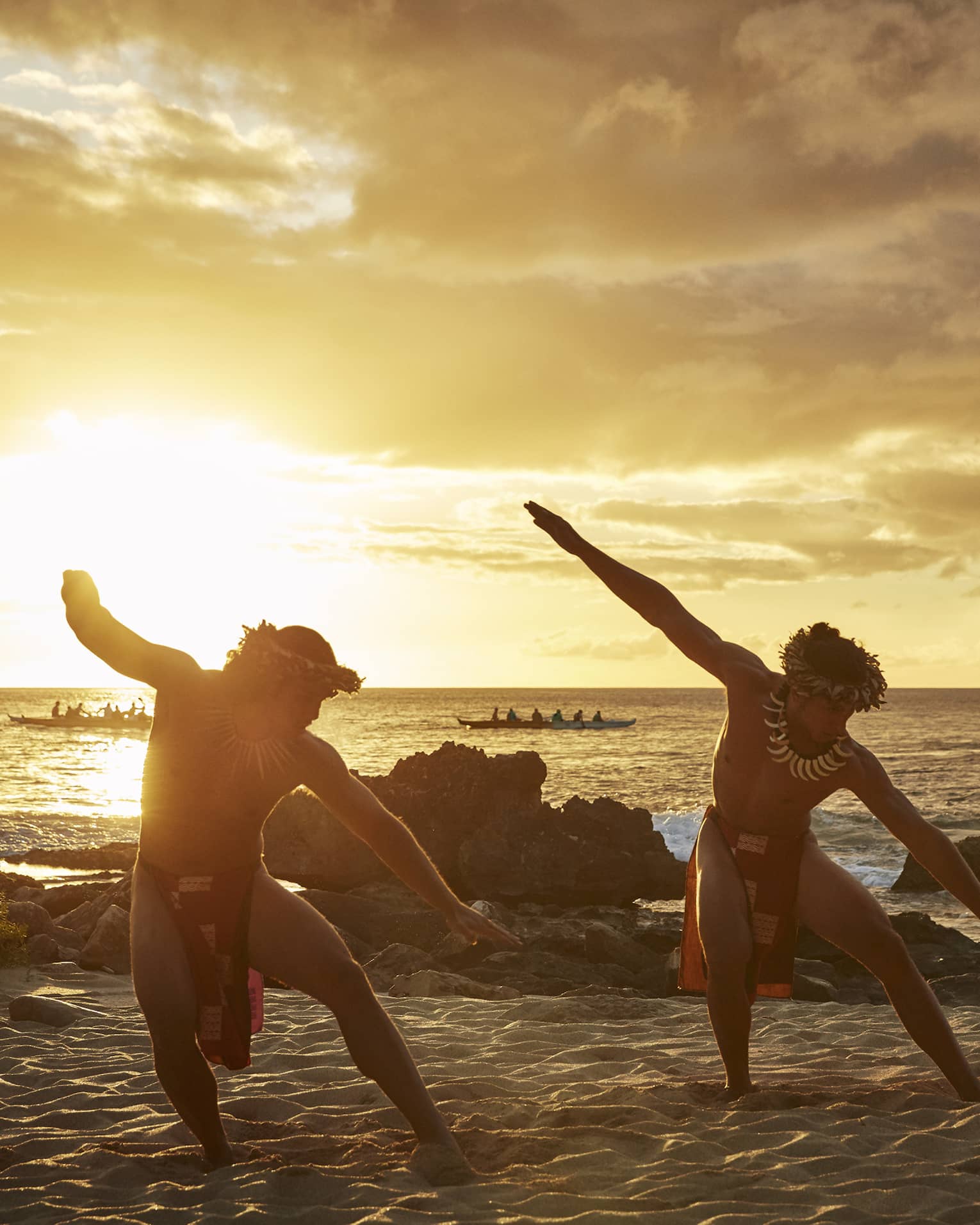 Two Hawaiian dancers with arms outstretched pose on sandy beach at sunset