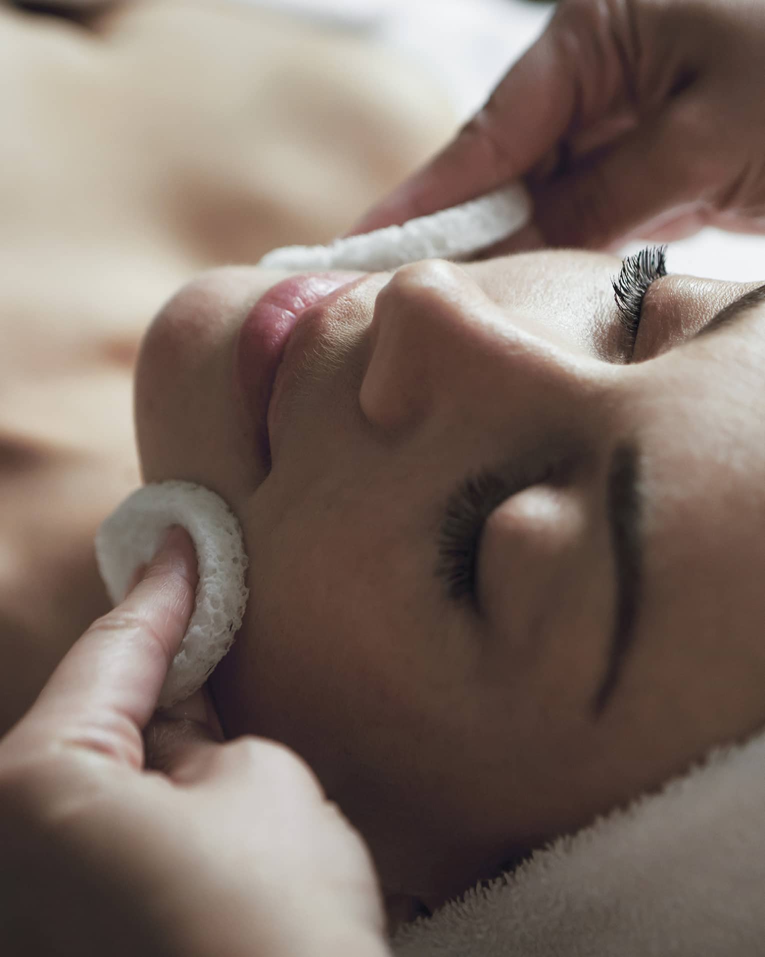 A detail of hands rubbing a sponge on a woman's face as she lies on a massage table with her eyes closed and hair wrapped up in a white towel
