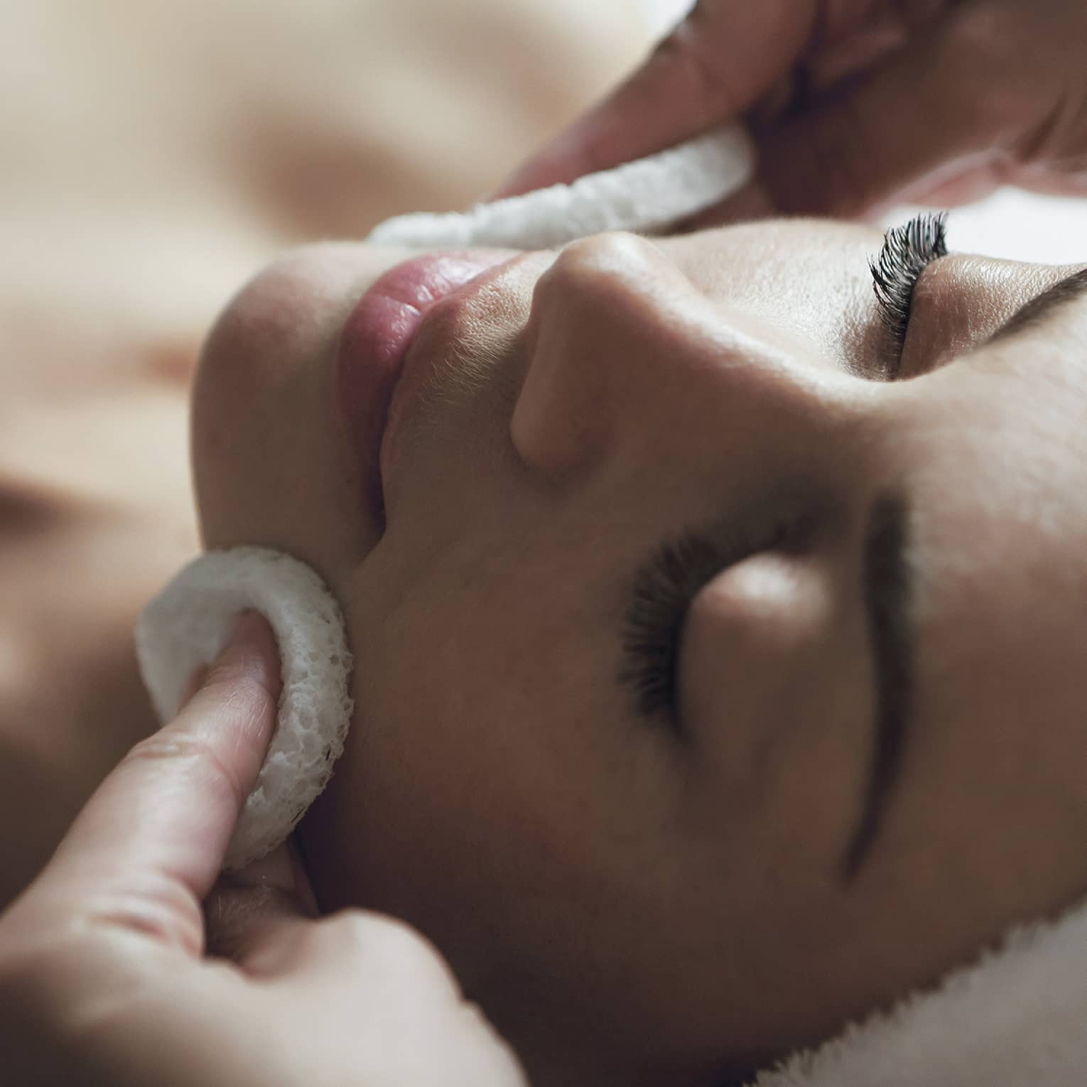 A detail of hands rubbing a sponge on a woman's face as she lies on a massage table with her eyes closed and hair wrapped up in a white towel