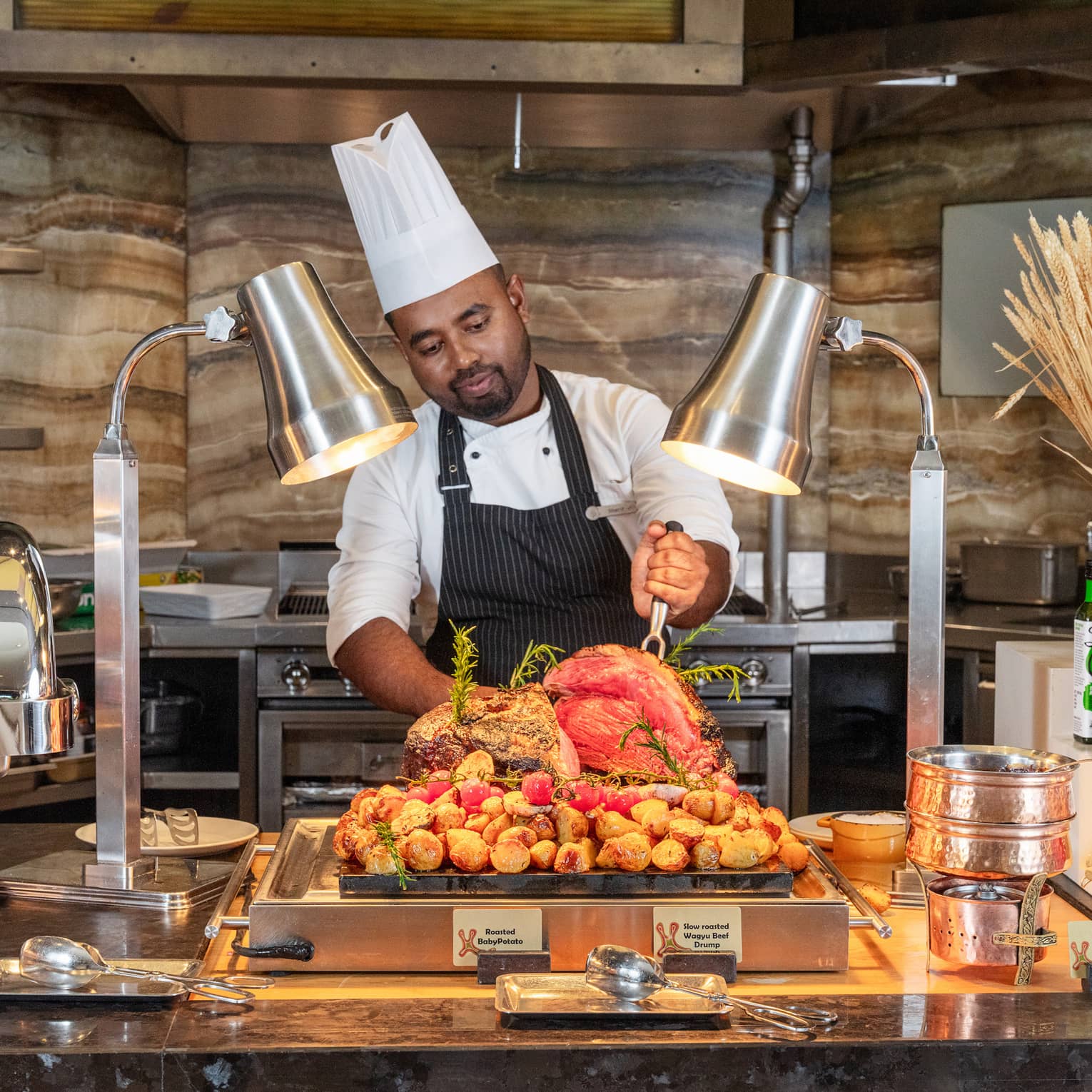 Chefs wearing tall white hat, white shirt and dark blue apron prepares to carve meat at a buffet live station