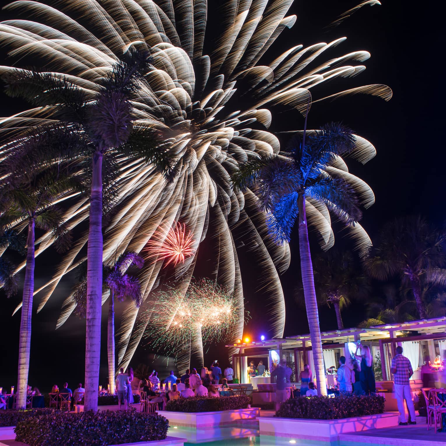 People around resort outdoor pool/bar watching fireworks in the night sky