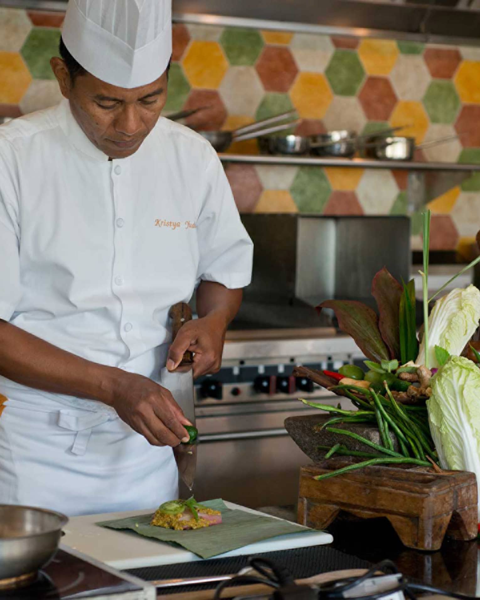 Chef in white uniform squeezes lime on large kitchen knife over dish, next to heads of fresh lettuce and whole peppers