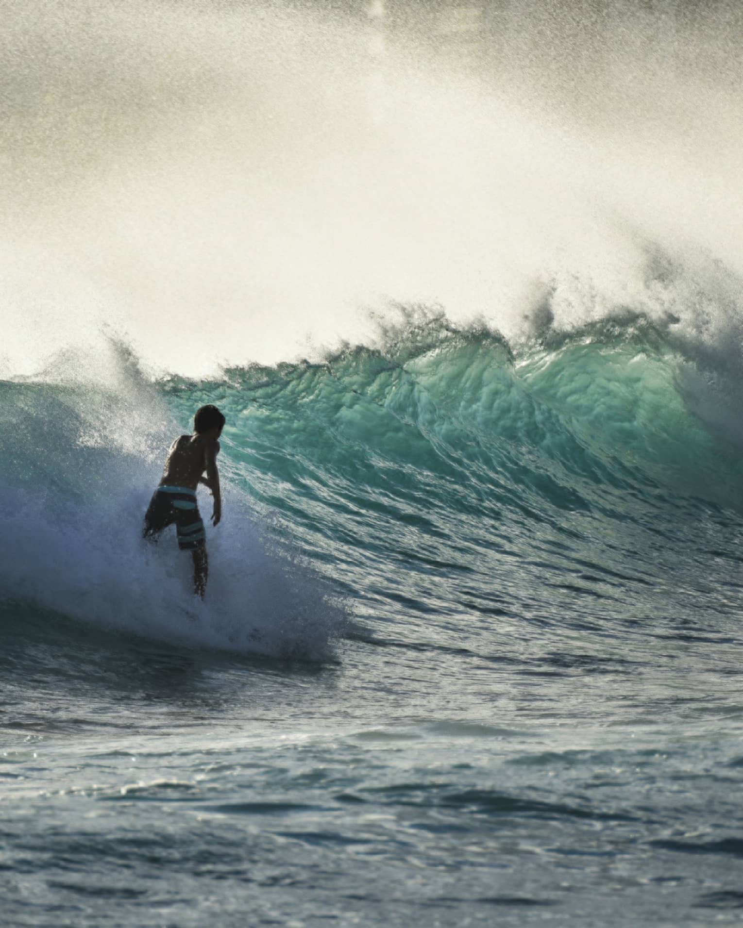 A surfer silhouetted in a spray of sea-foam rides a large wave in a teal-coloured ocean under a misty, yellow-streaked sky.