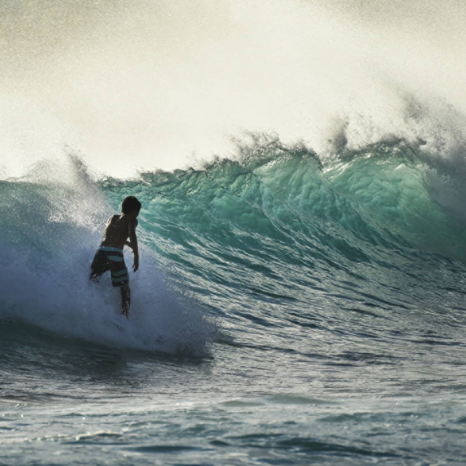 A surfer silhouetted in a spray of sea-foam rides a large wave in a teal-coloured ocean under a misty, yellow-streaked sky.
