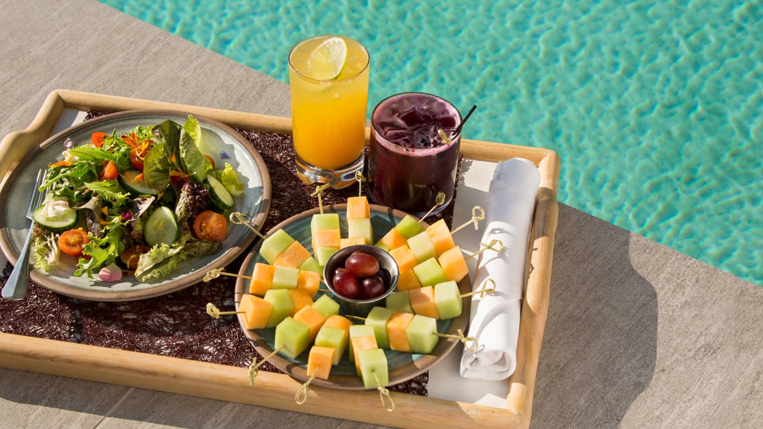 Tray with fresh fruit skewers, fresh salad, juices on tray beside swimming pool