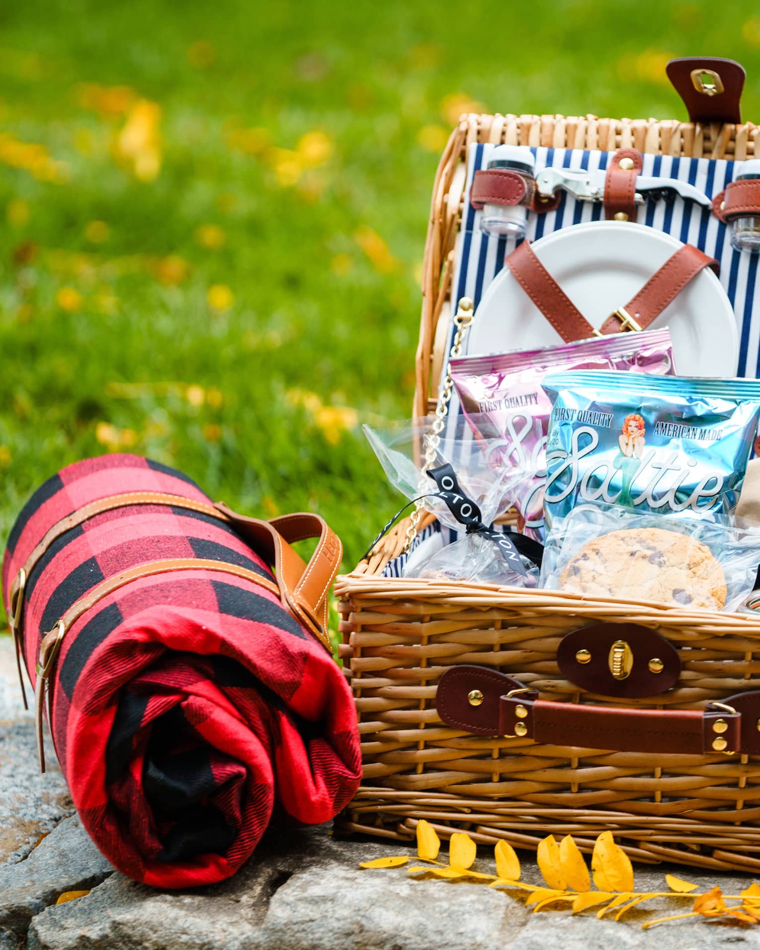 A picnic basket with plates, utensils and snacks resting on a stone near grass and yellow leaves.