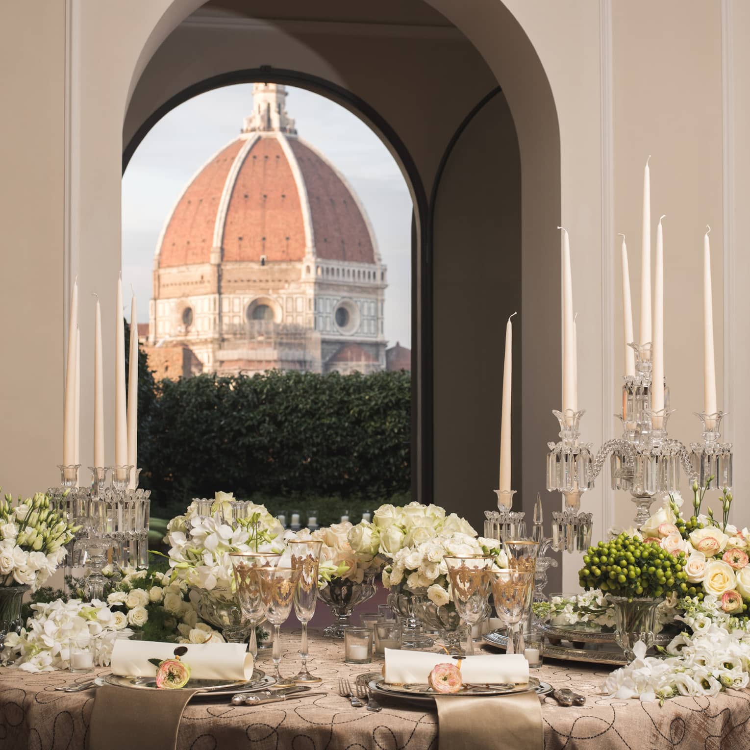 Elegant dining table set with candles, flowers, and glassware, with a large dome visible through arched windows in the background.