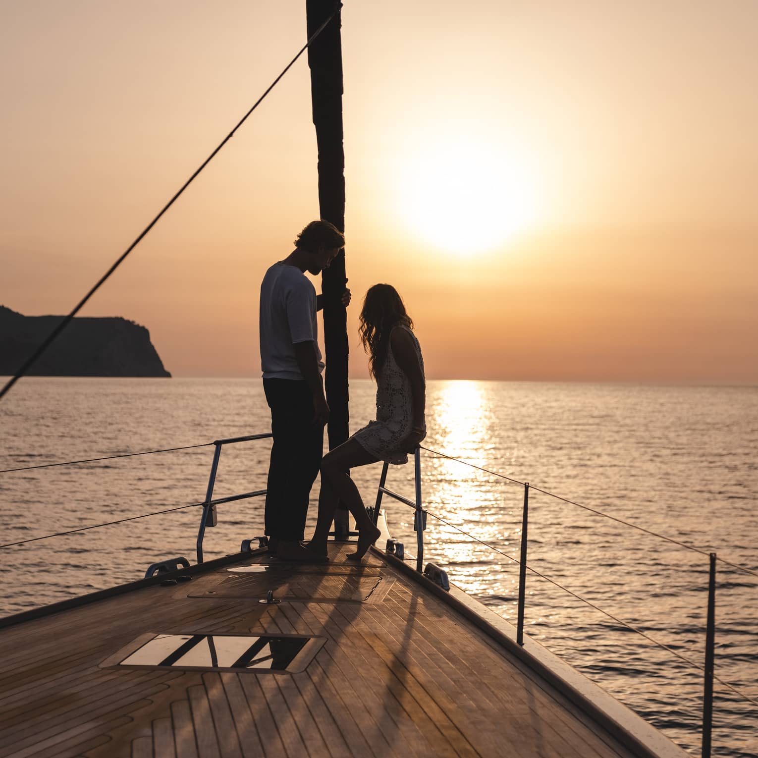 A couple at the pointed bow of a boat is silhouetted against the warm glow of the sun setting over calm, rippling water.