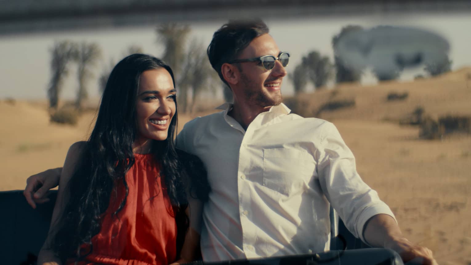 A couple dressed nicely sitting in a car with the dessert behind them.