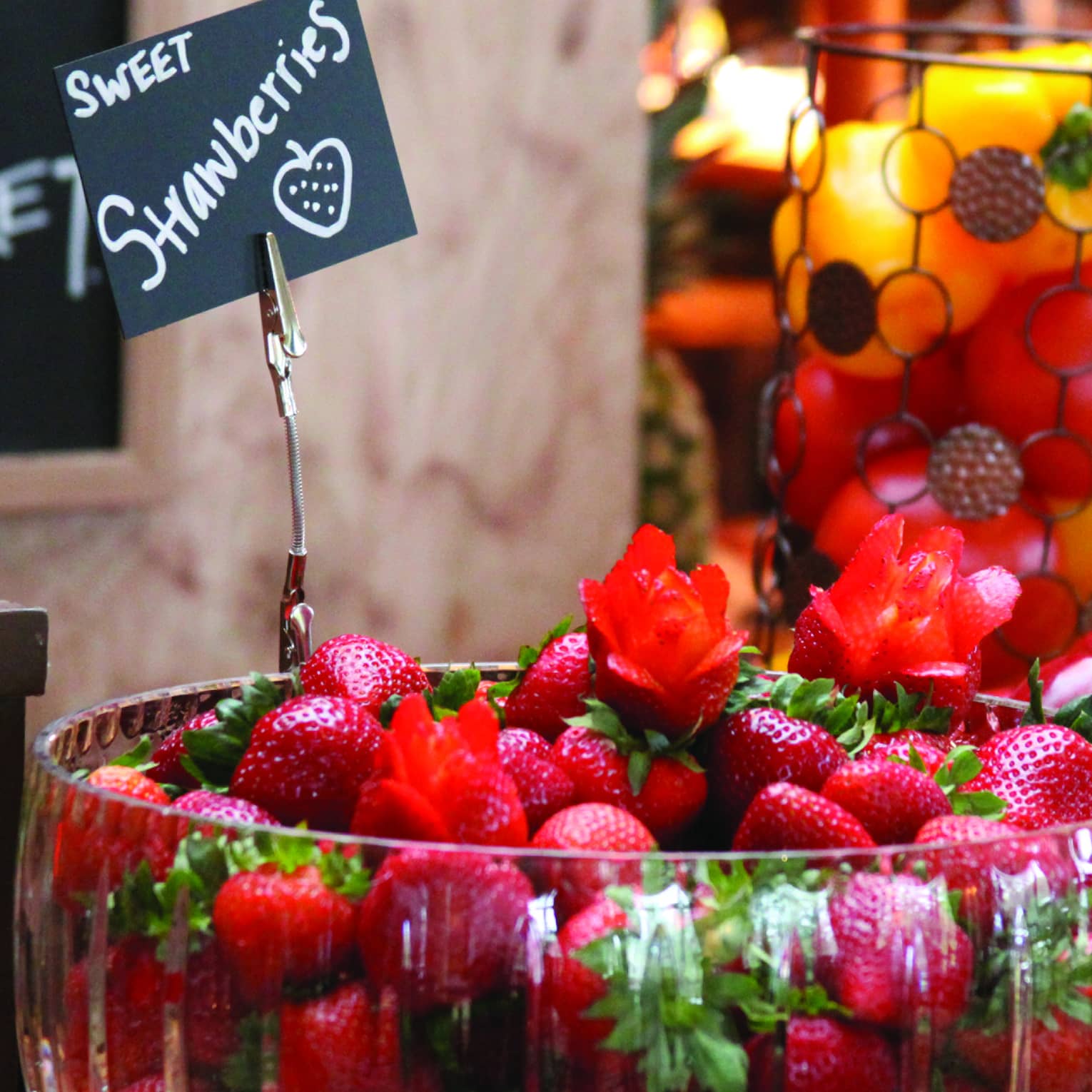 A table in a market displaying a glass bowl of fresh strawberries and an assortment of apples, peaches and peppers.