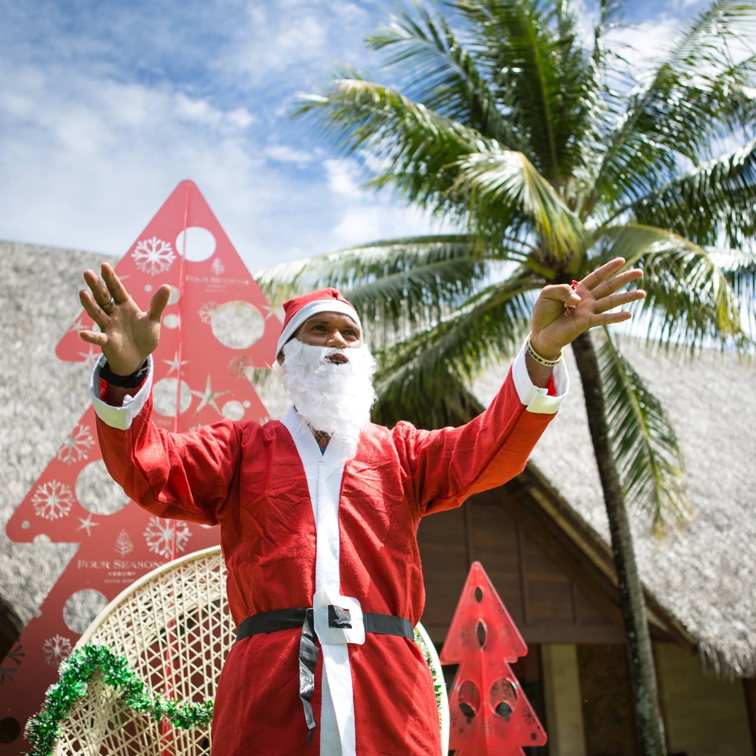 Person in Santa suit stands outside grass-roof structure with arms outstretched