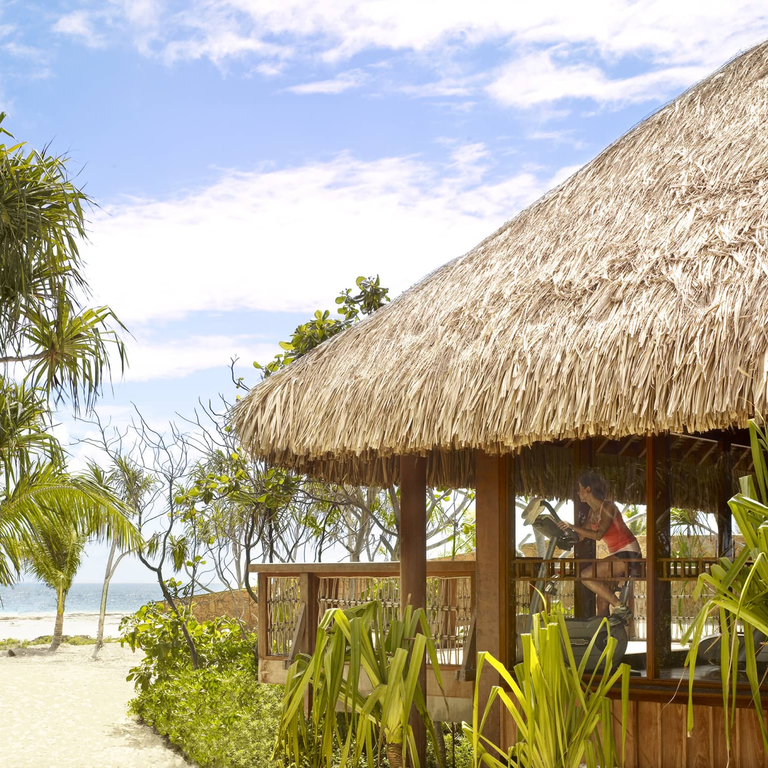 Woman on exercise bike in thatched-roof gym looks out at beach