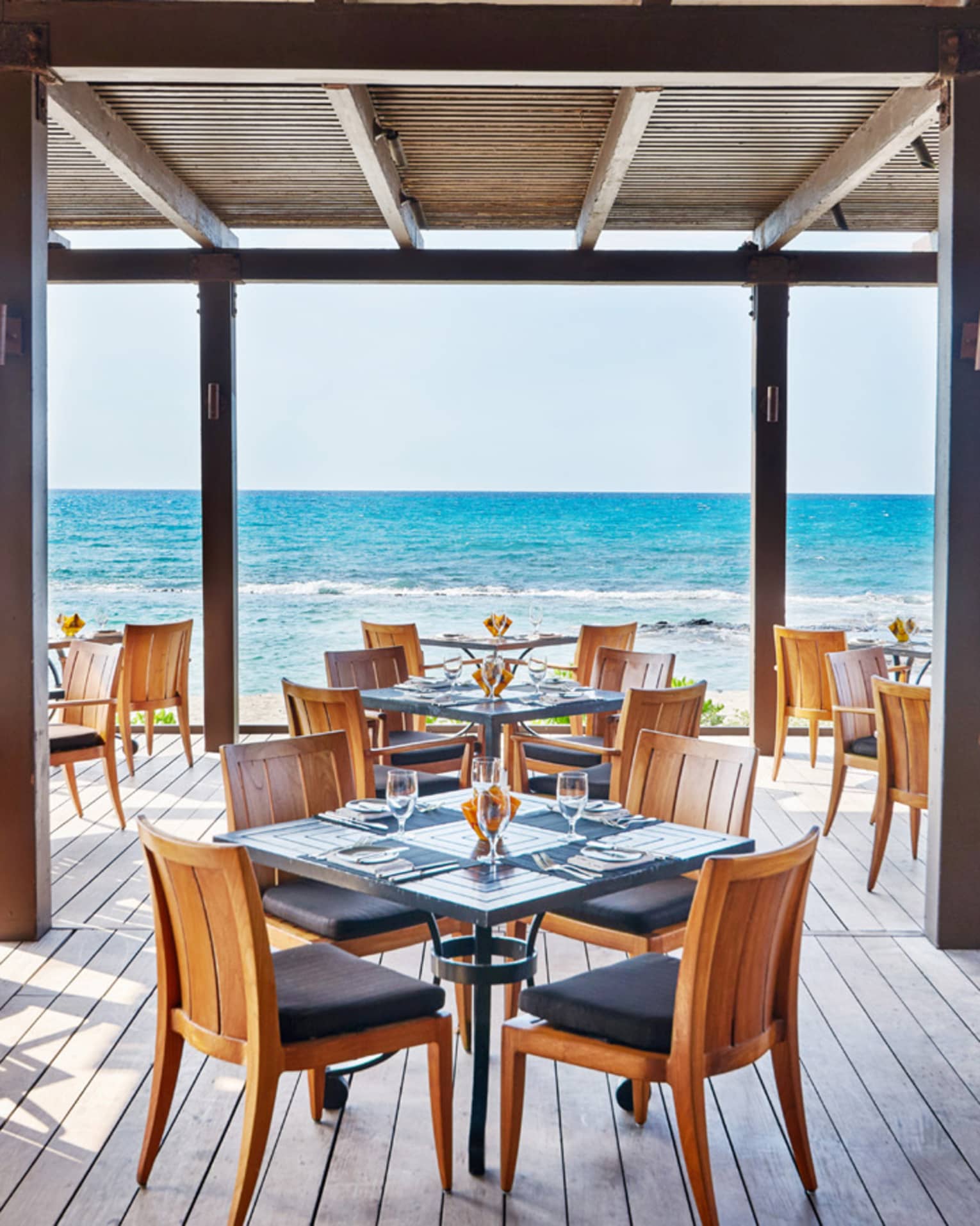  The Ulu Ocean Grill and Sushi Lounge is ready for guests with square tables set with menus and clear glassware, and overlooking the ocean from the covered wooden terrace. 