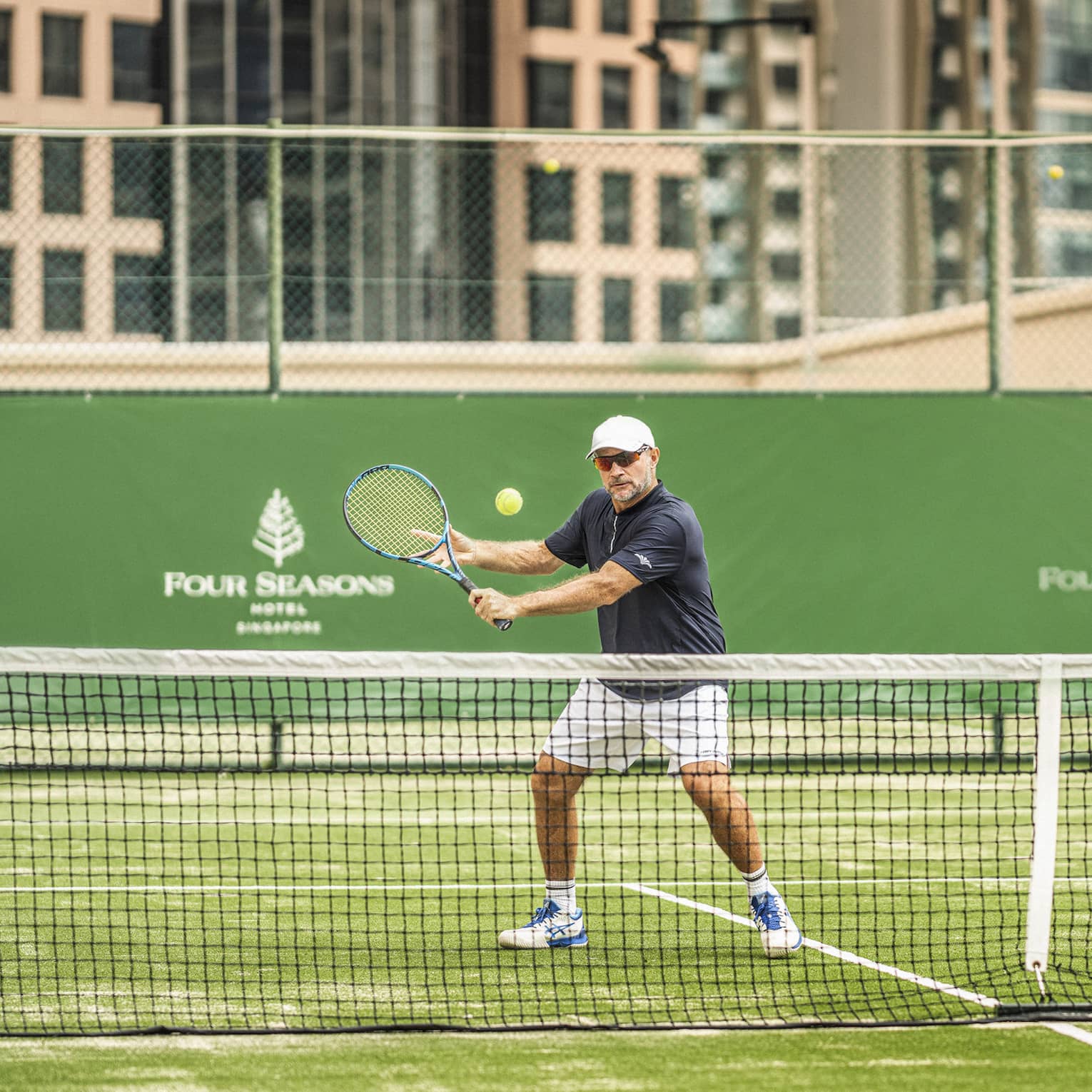 Under a dusty-pink sky with tall buildings looming behind, a tennis player holds a racquet aloft as a ball approaches.