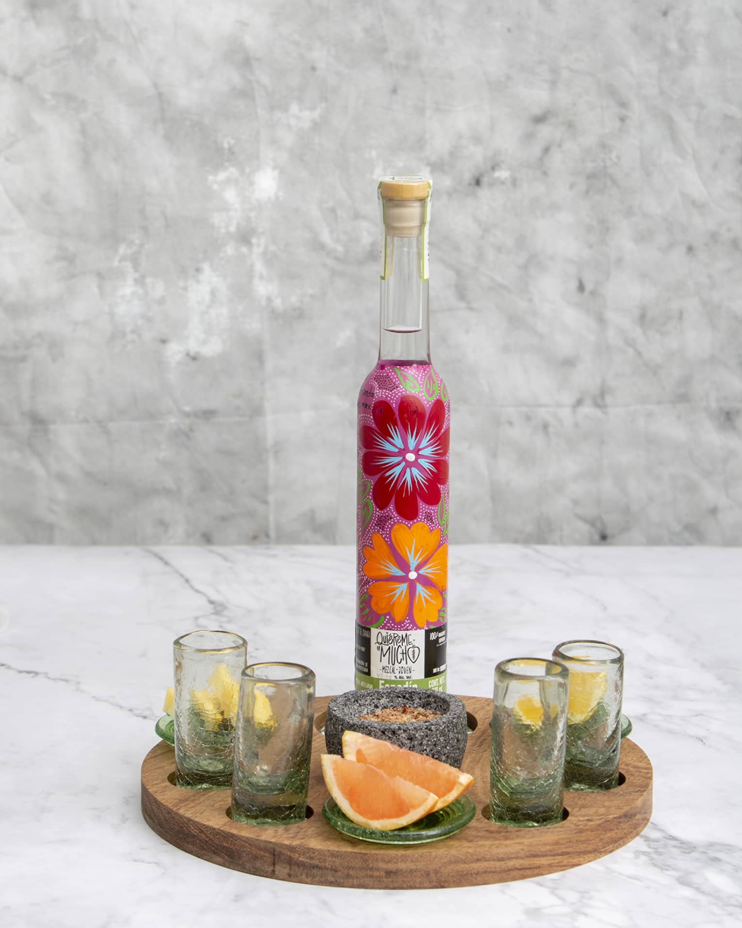 Brightly floral-painted mezcal bottle on wooden tray with four shotglasses, stone spice bowl and lemon and grapefruit wedges.