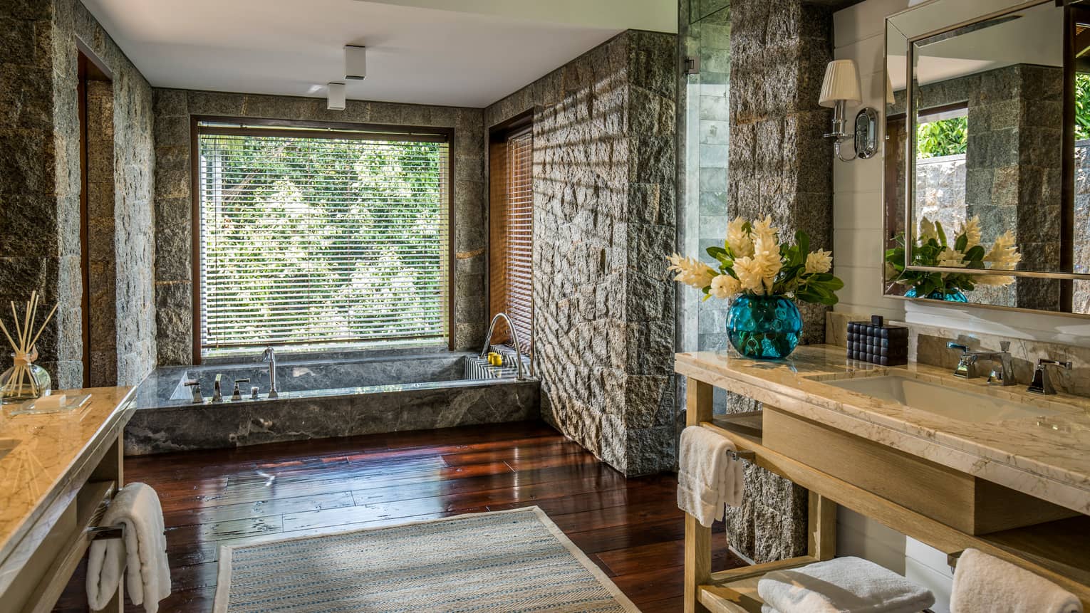 Long vanities in large bathroom with stone walls, sunken marble tub under window