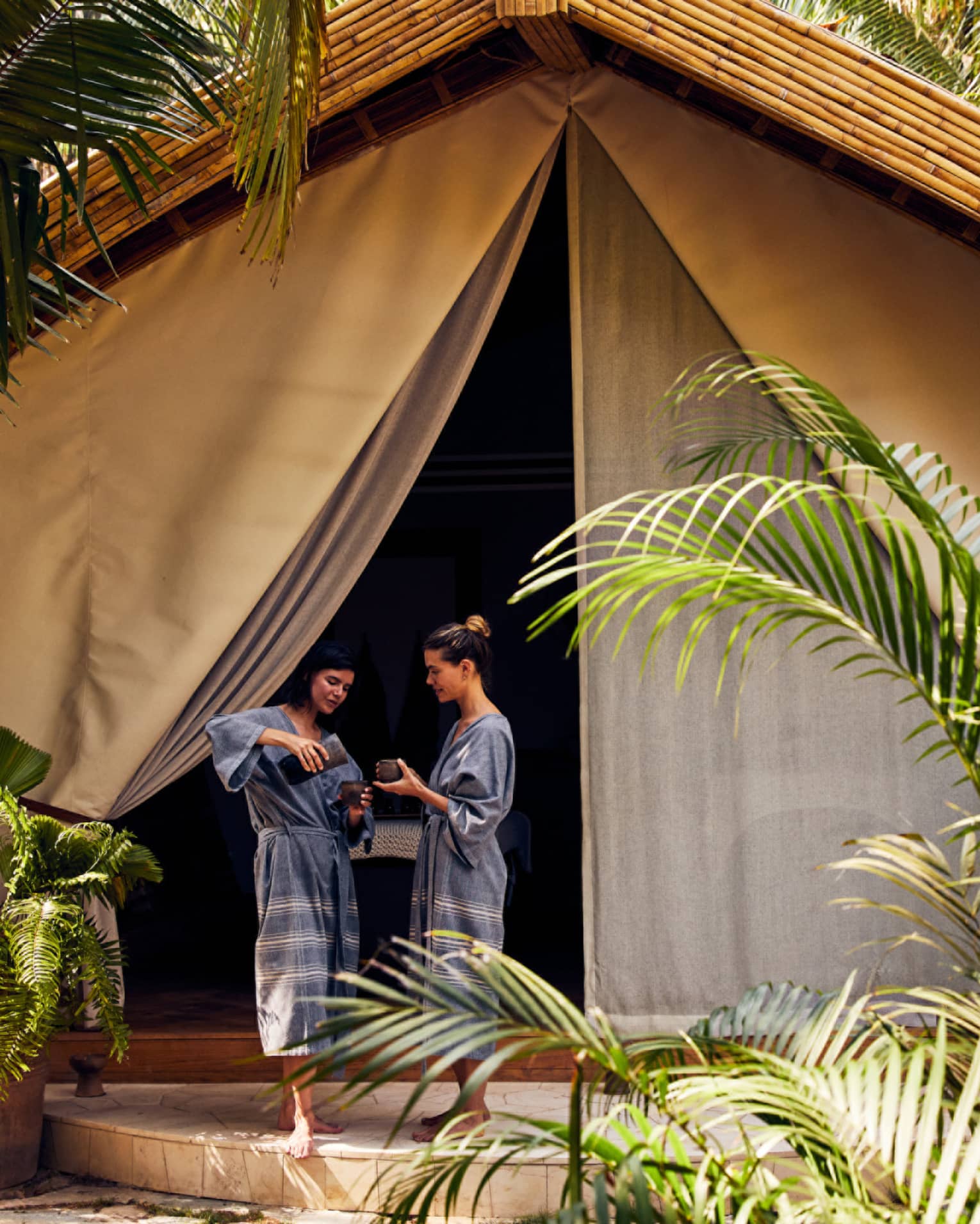 Two women in spa robes entering a spa tent surrounded by palm trees.