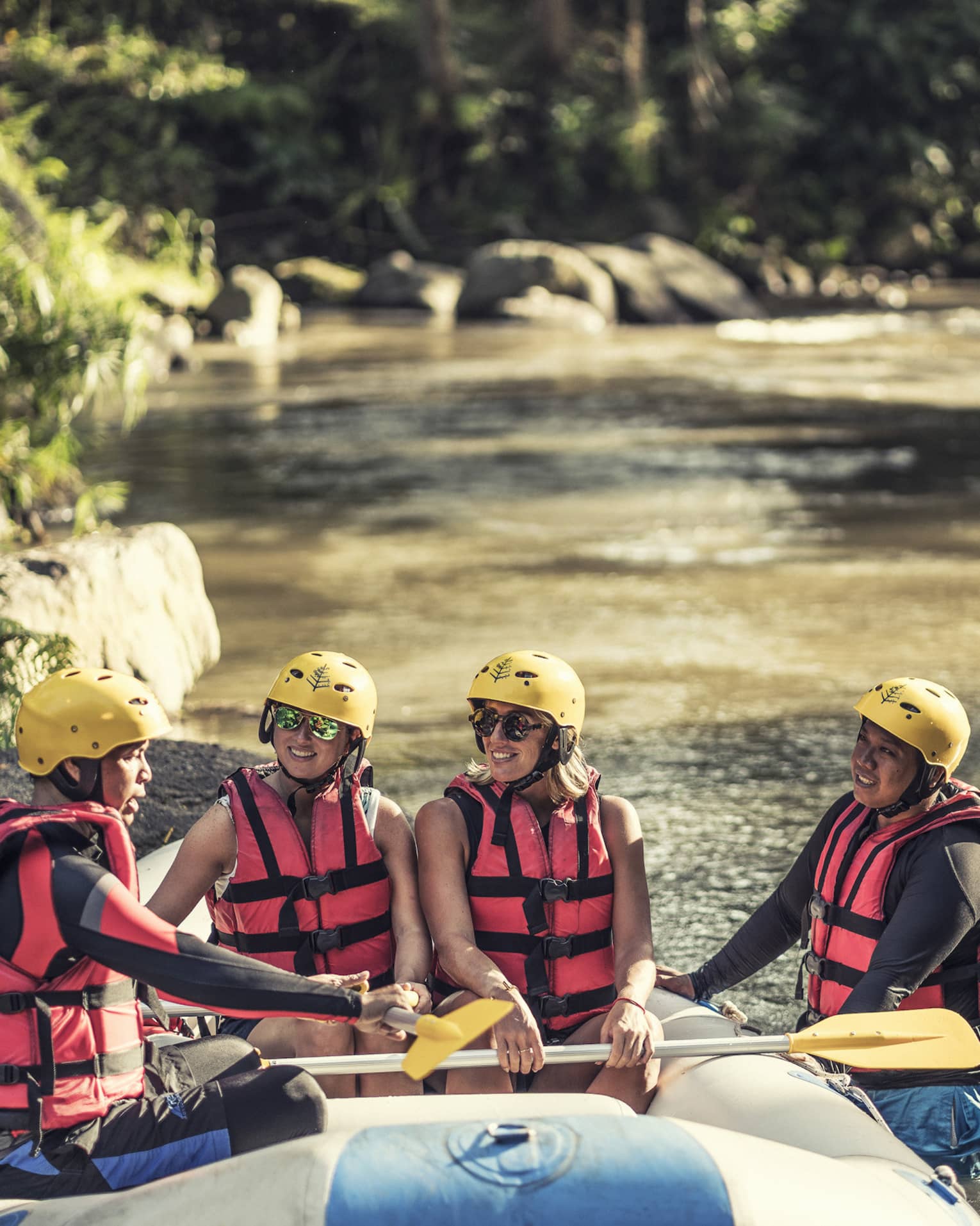 Four people wearing red life jackets, sunglasses and yellow helmets sit facing each other in a river raft