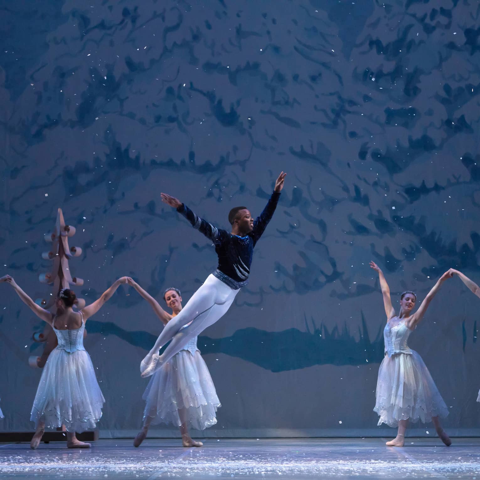 Ballerinas on stage in long ice-blue tutus dance before a snowy painted backdrop as a dancer wearing a leotard leaps grandly.