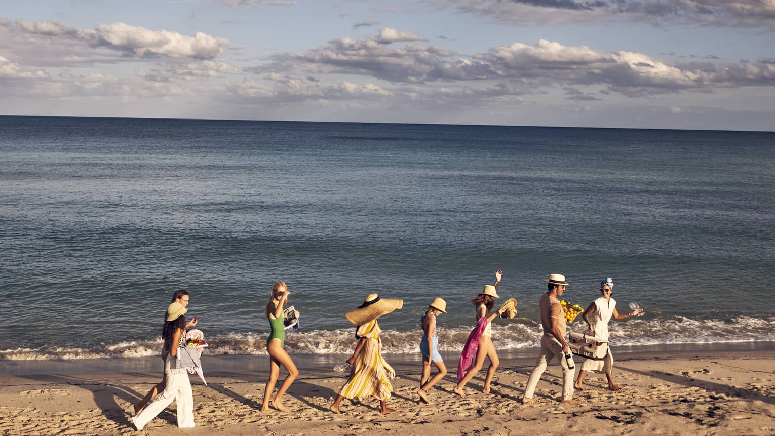 A group of people walking along a sandy beach by the ocean, wearing summer clothing and hats, with a backdrop of a calm sea and partly cloudy sky.