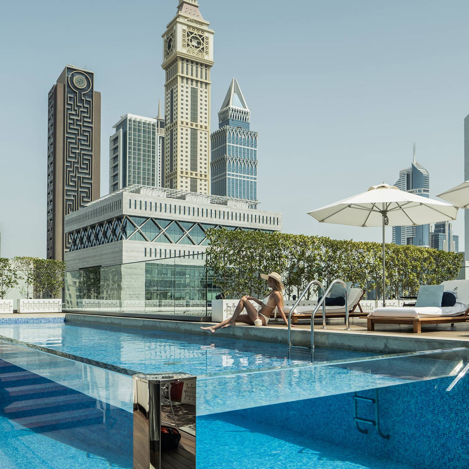 A guest lounges by a transparent rectangular rooftop pool, modern towers with geometric designs rising into the azure sky.