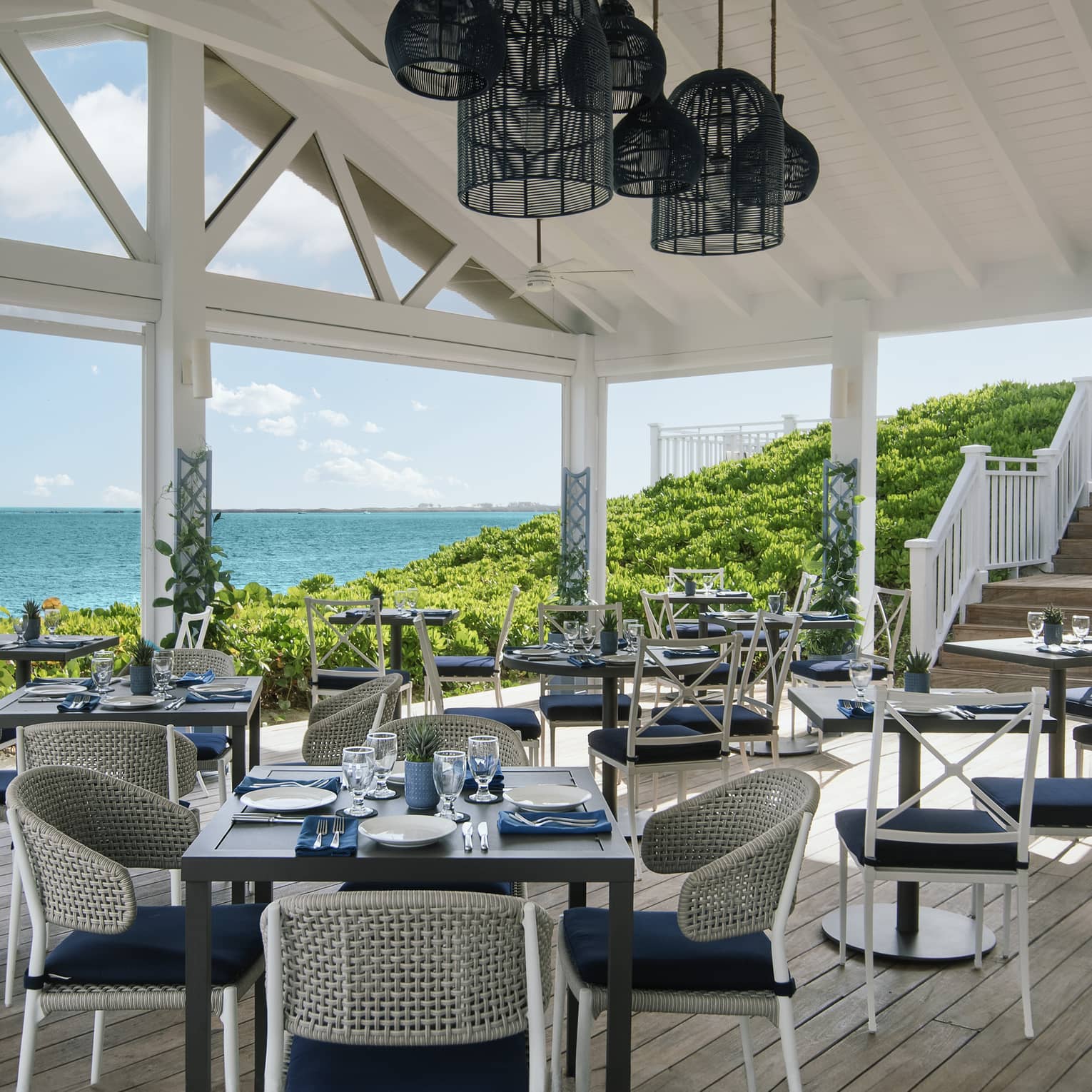 Covered restaurant patio with vasious square tables each set with four blue-and-white chairs with a view of the ocean in the background